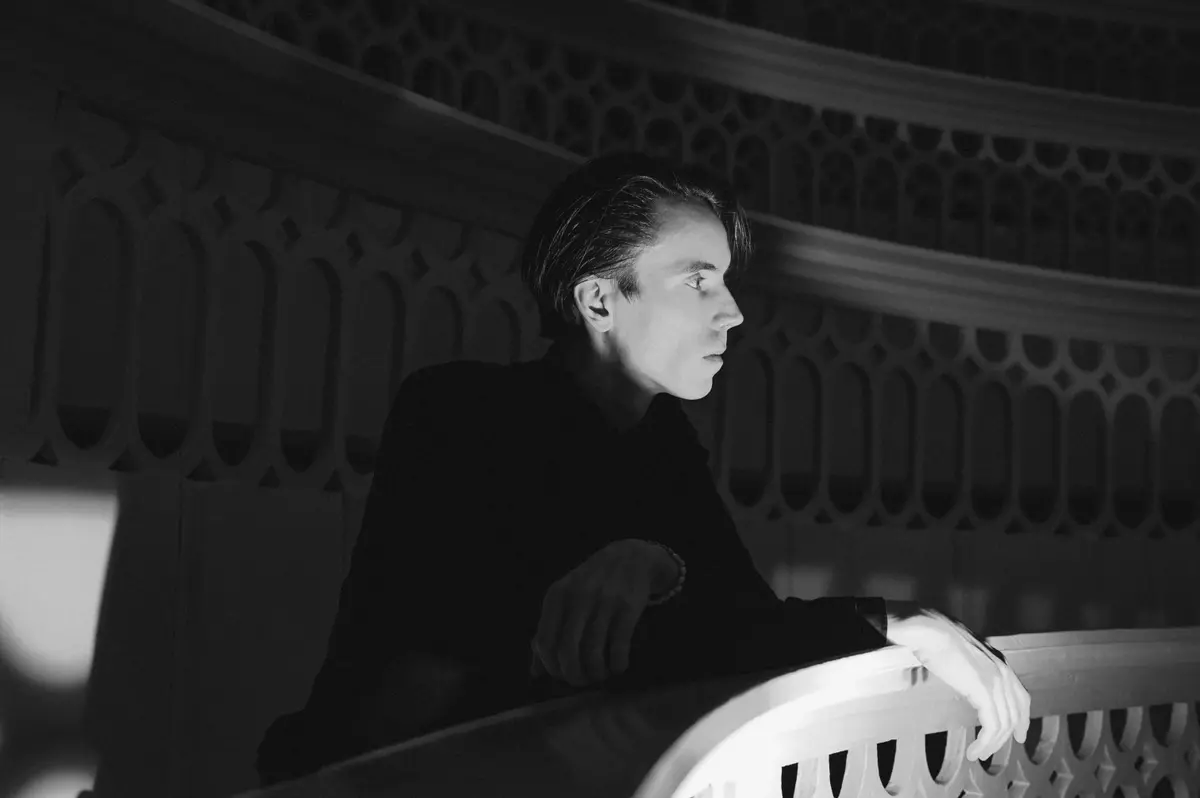 A person stands in a dimly lit area, leaning on a railing with a patterned background.