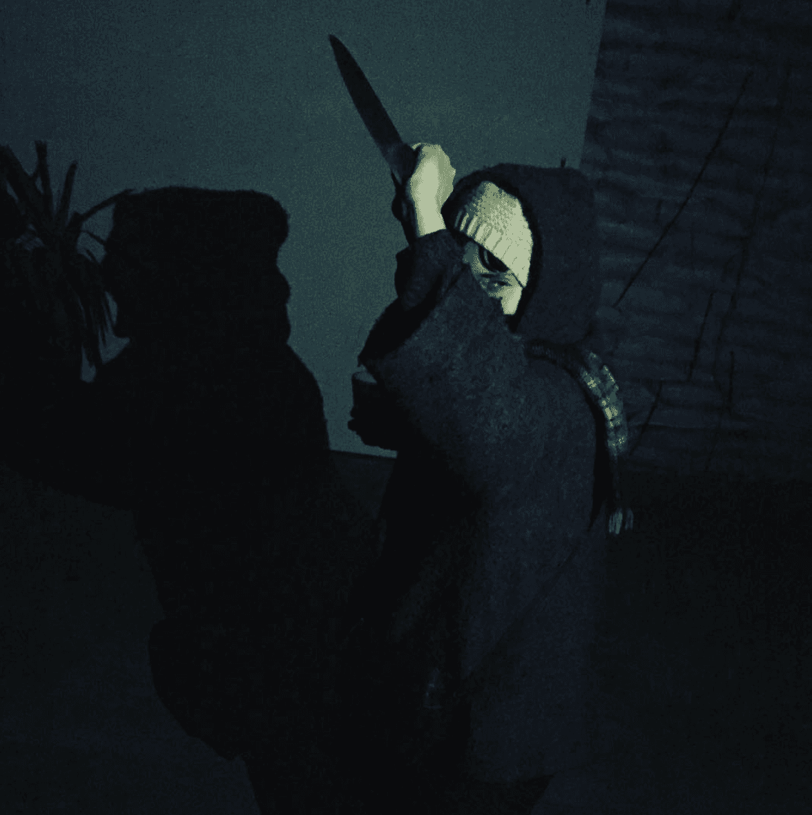 A person in a hooded sweater holds a knife aloft in a dimly lit setting, casting a shadow on the wall.