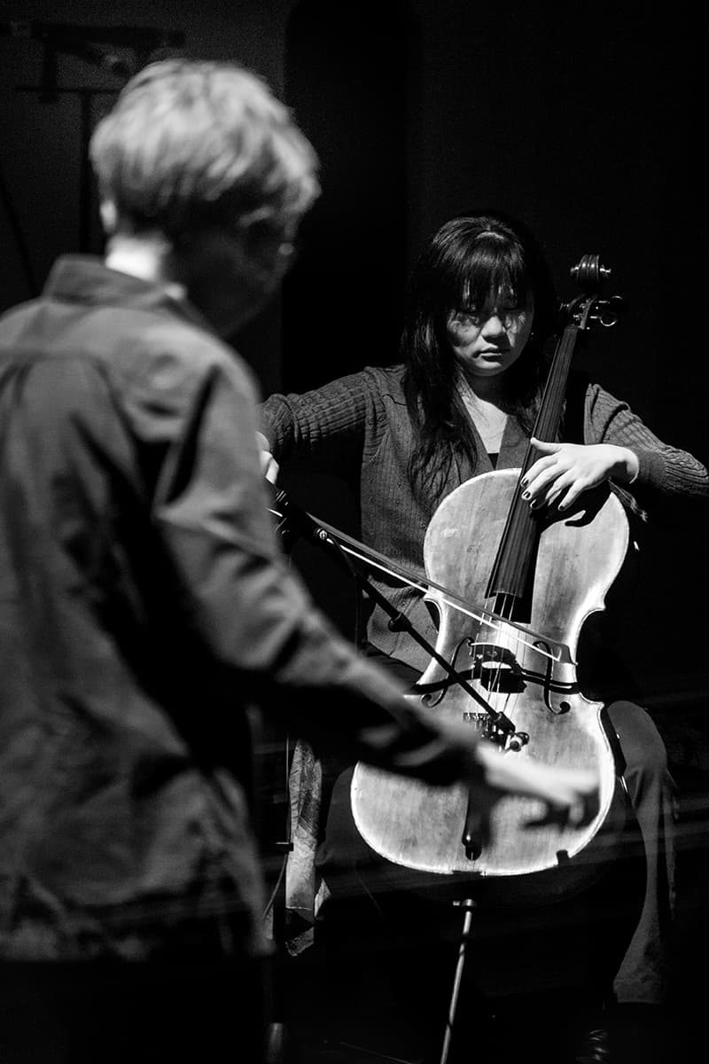 A woman plays the cello, while another person stands in the foreground, in a black and white setting.
