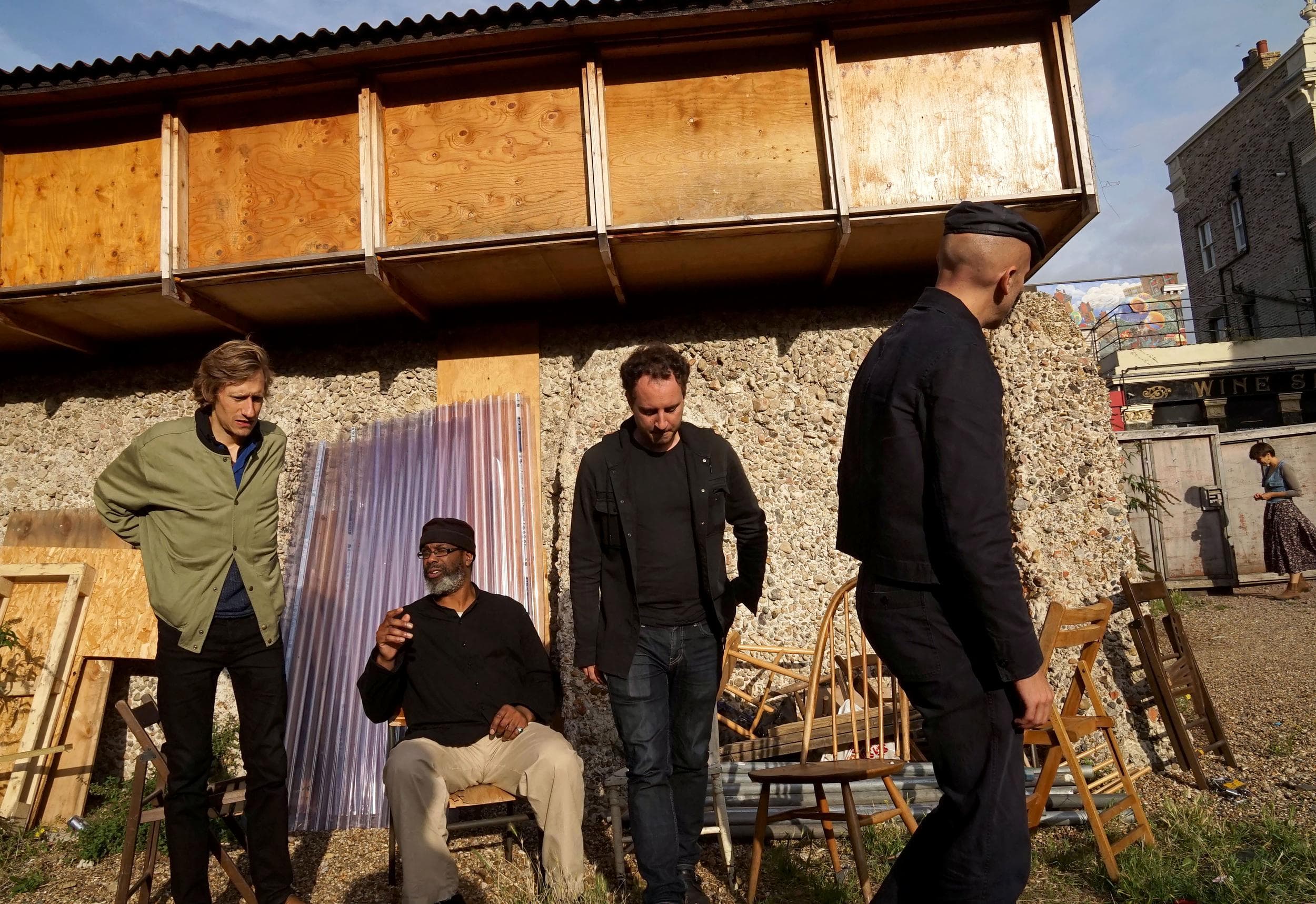 Four people are gathered outside near a textured wall and wooden structures, with chairs and building materials around them.