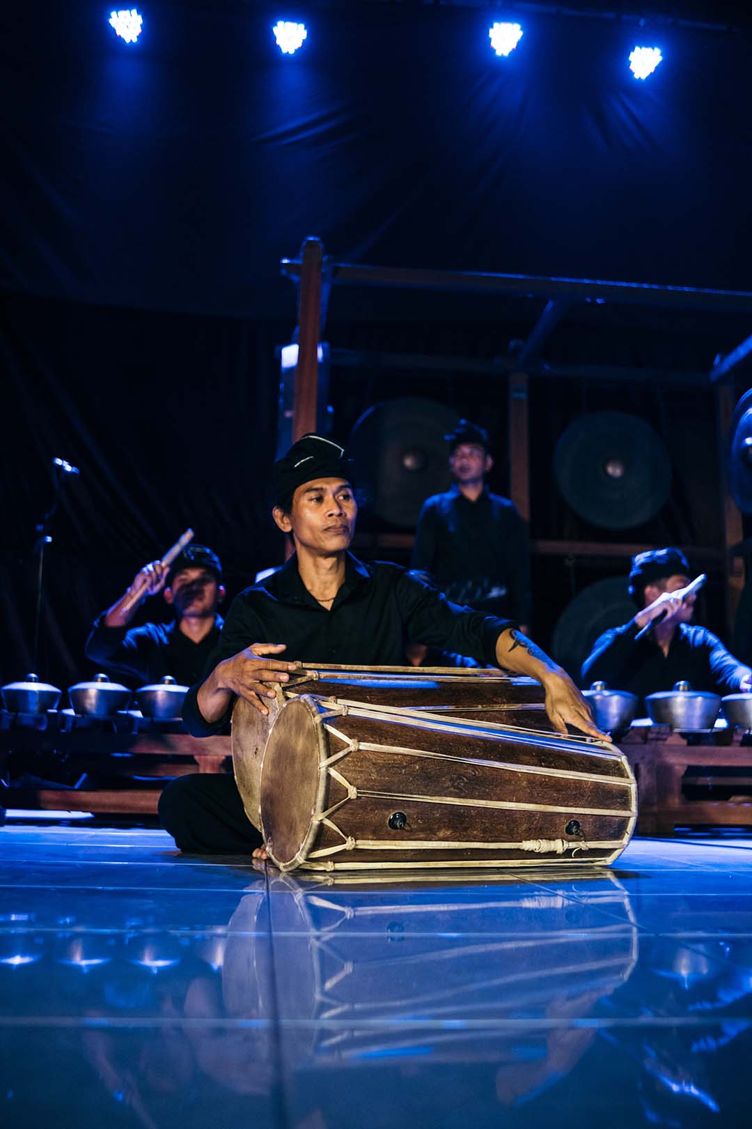 A musician is seated on stage playing a large traditional drum, with other musicians and instruments in the background under blue lighting.