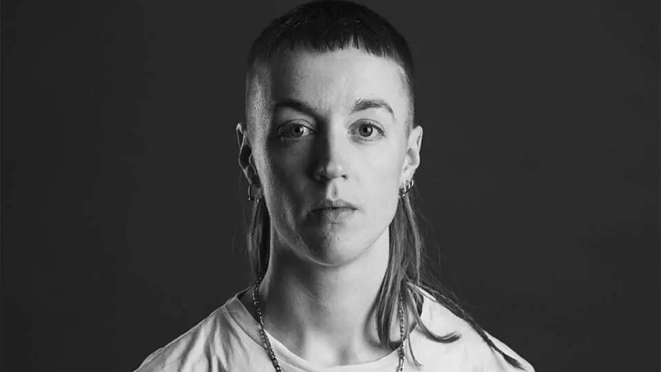 A person with a mullet hairstyle and earrings is facing forward against a dark background.