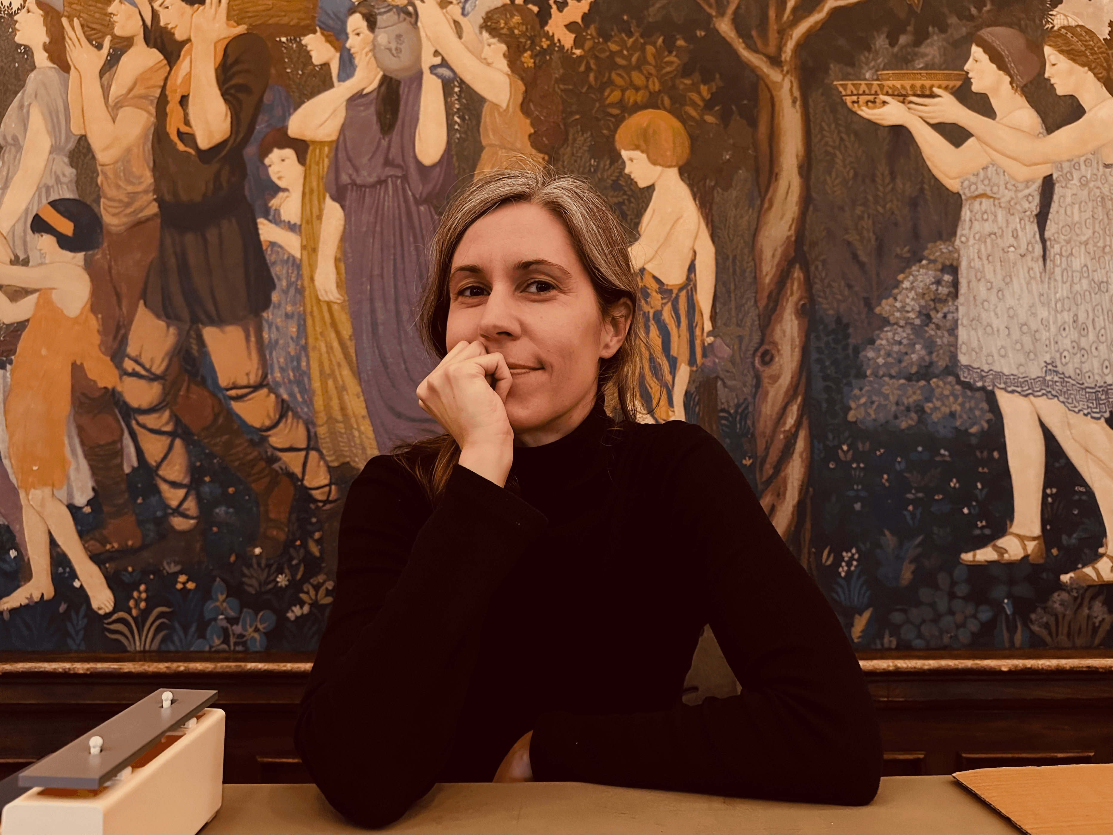 A woman with light hair and a black turtleneck sits at a table with her chin resting on her hand, in front of a colorful mural depicting people.