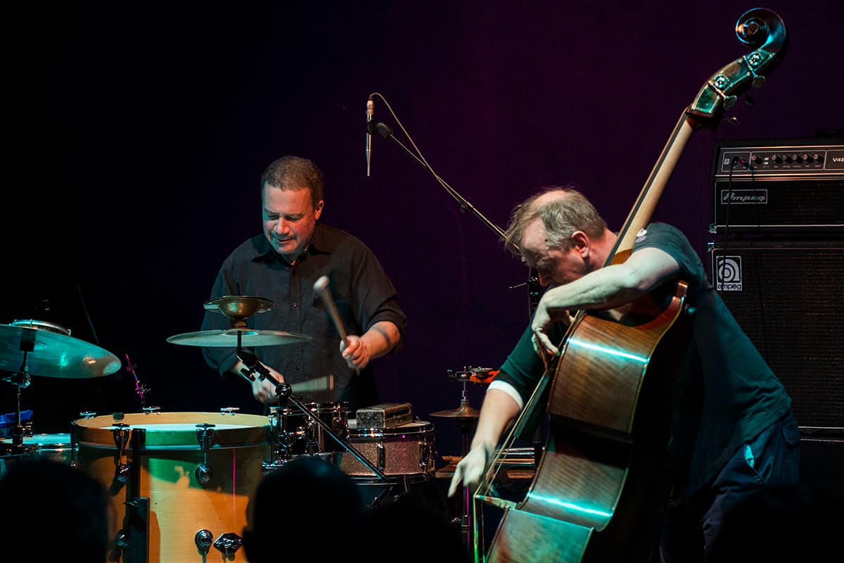 Two musicians perform on stage, one playing drums and the other playing an upright bass.