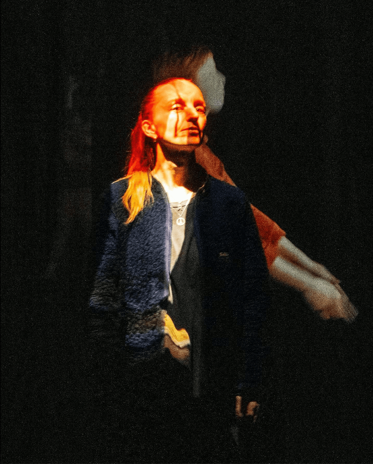 A person stands in front of a dark background with a colorful light projection on their face and body.