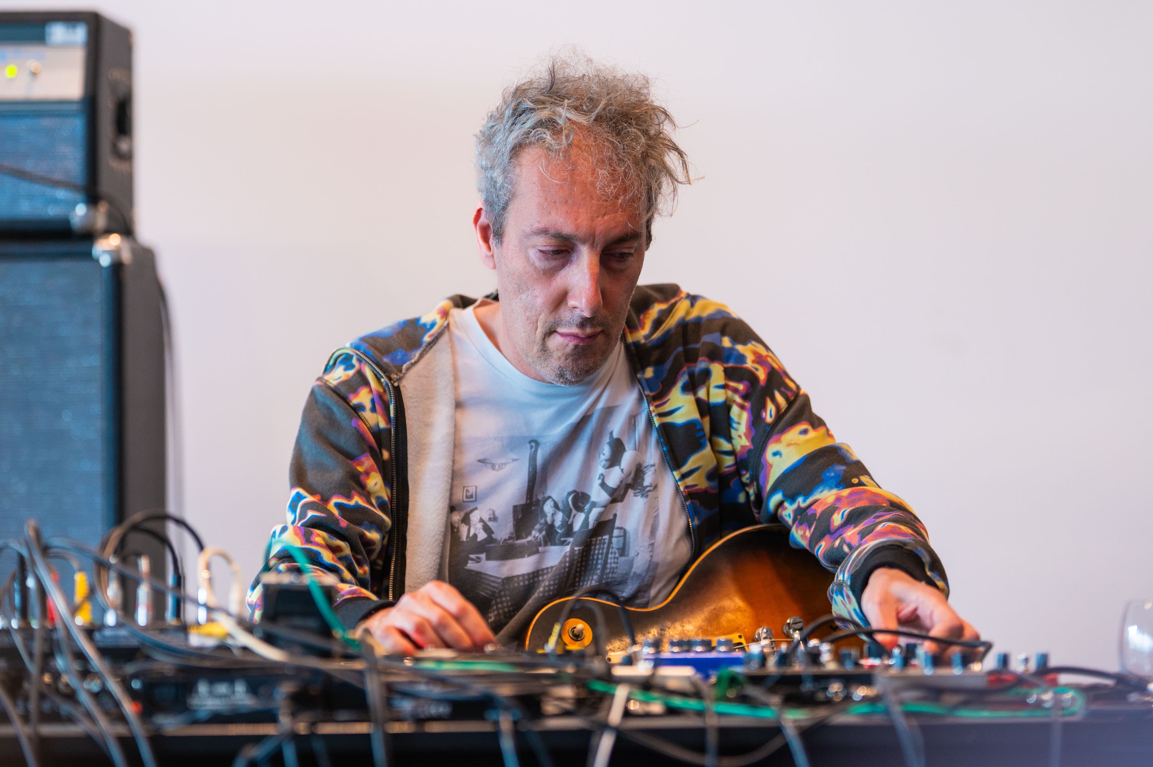 A person with gray hair is adjusting audio equipment, with a guitar partially visible and wearing a colorful jacket.