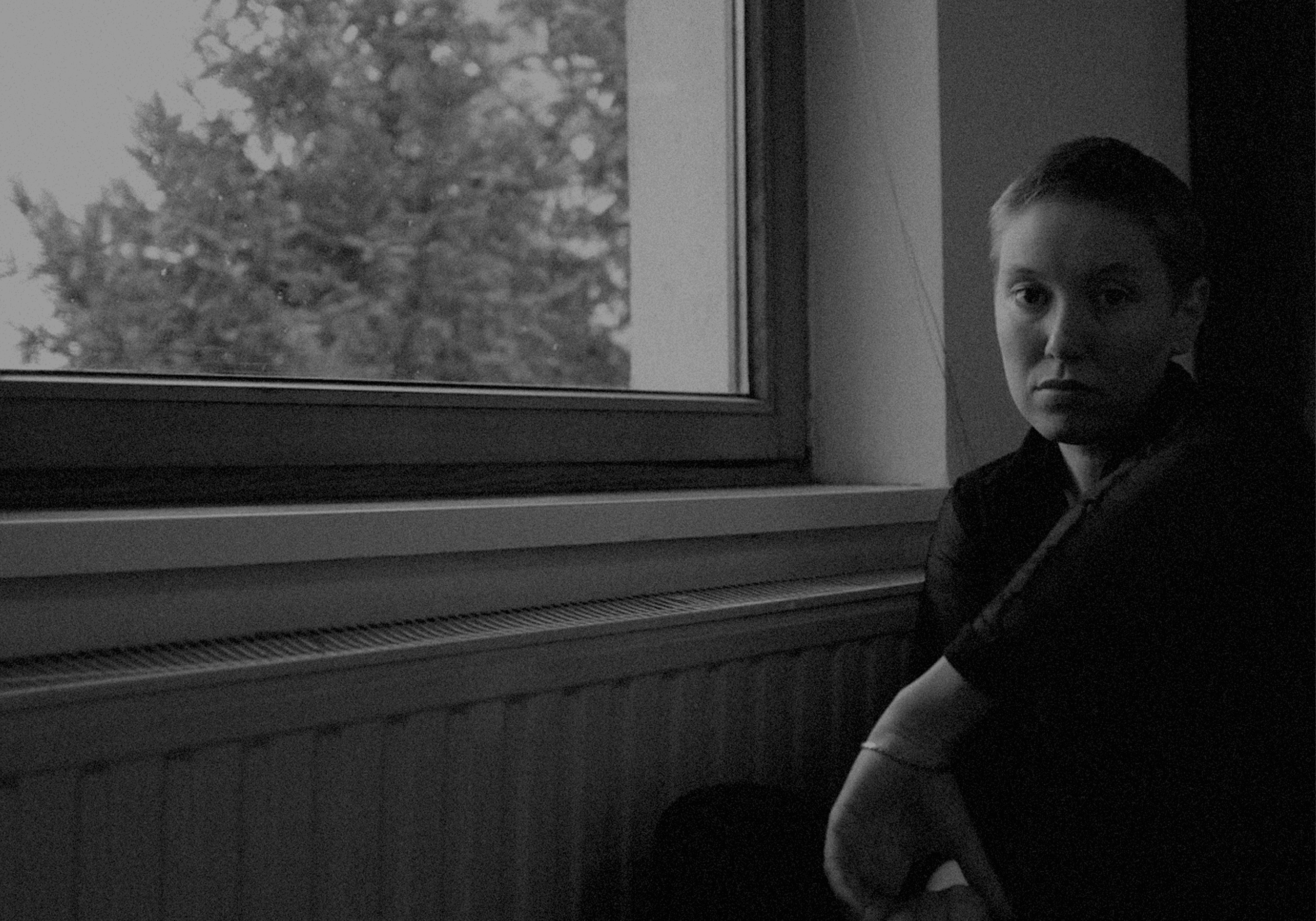 A person with short hair sits by a window, looking towards the camera, in a dimly lit room.