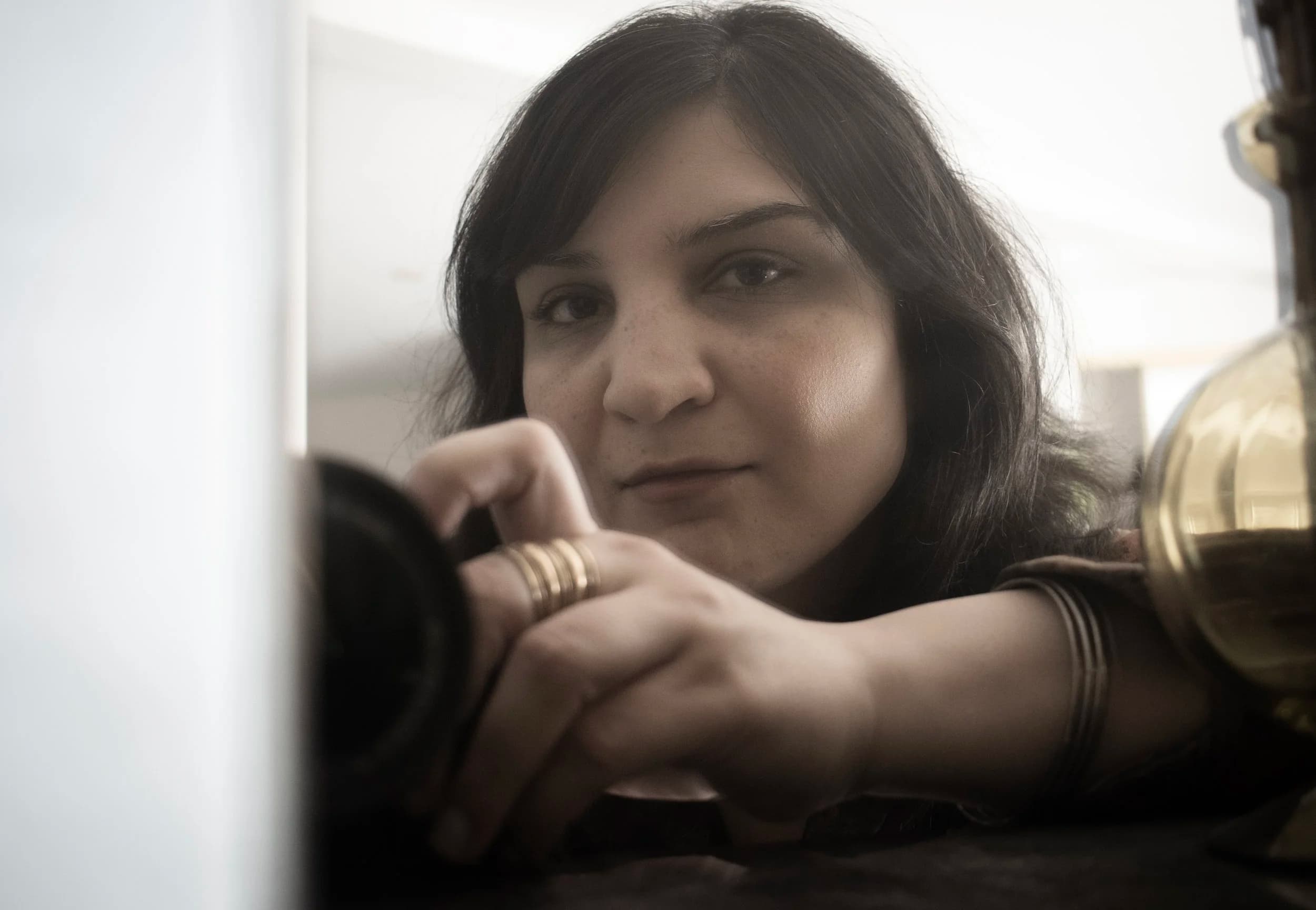 A person with long dark hair leans forward, holding a camera with one hand, wearing rings and a bracelet.