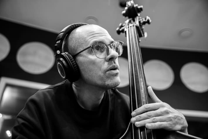 Black and White photograph of man playing a double bass. He has a shaved head and wears both glasses and headphones. He is leaning forward with his left hand over the strings. Wall in background has a row of circles.