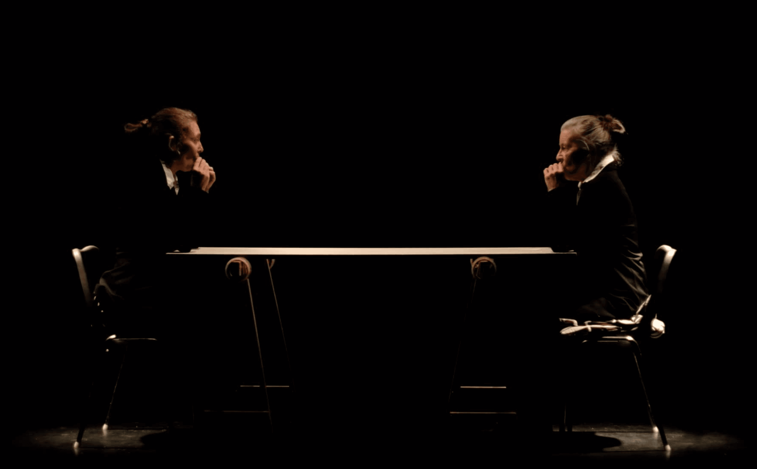Two people dressed in dark clothing sit facing each other at a table in a dimly lit setting.