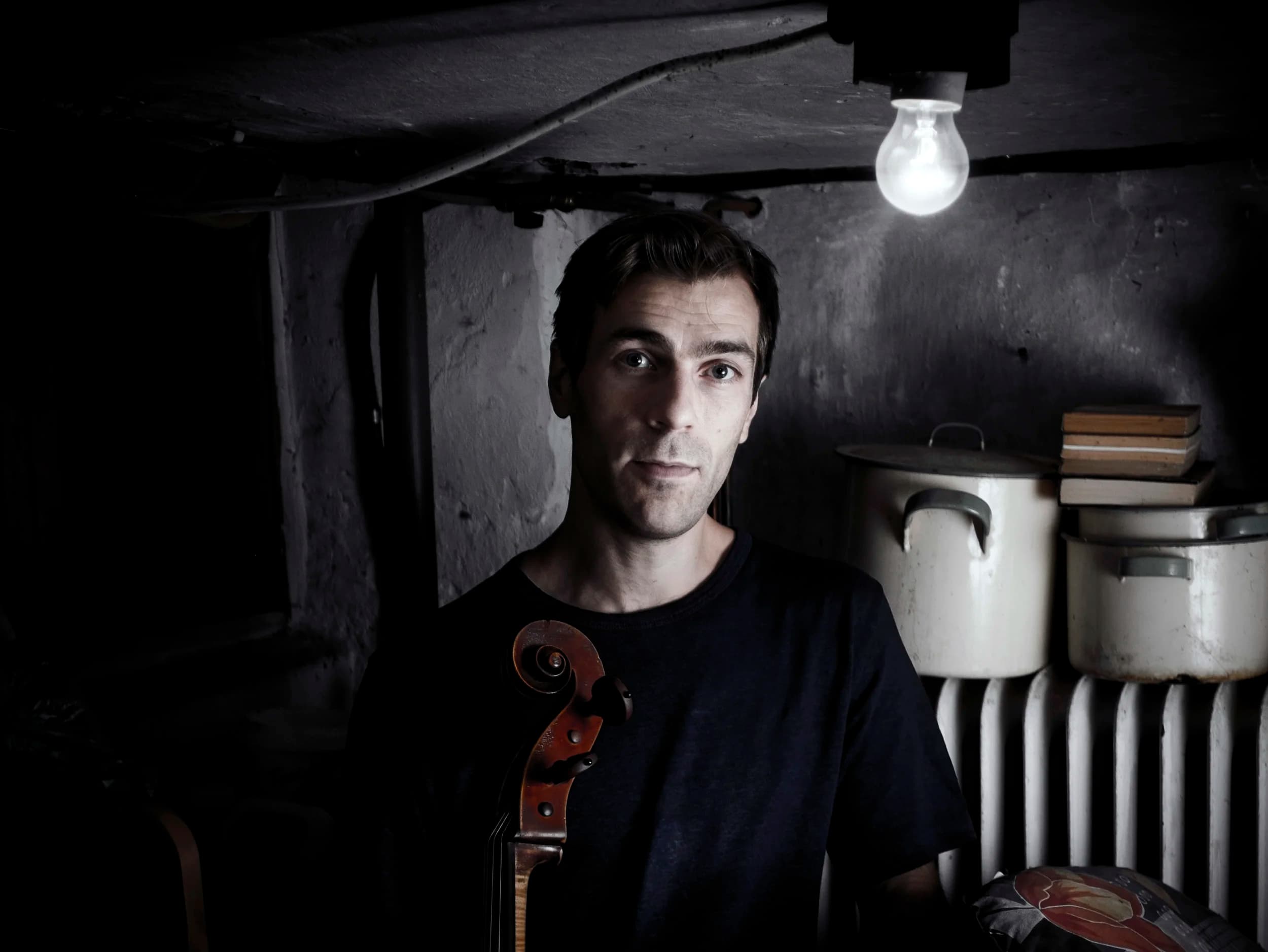 A man stands in a dimly lit room holding a cello, with a light bulb overhead and pots stacked nearby.