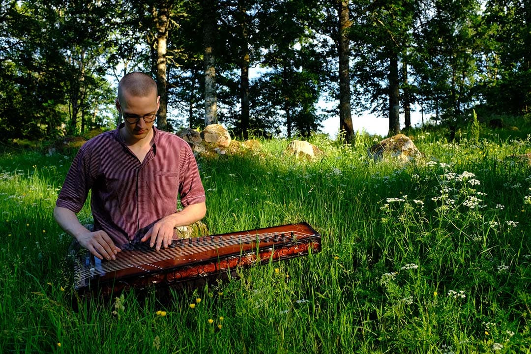 A person sits in a grassy field playing a stringed instrument with trees in the background.