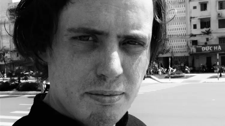 A person with medium-length hair looks at the camera, with buildings and a street in the background.