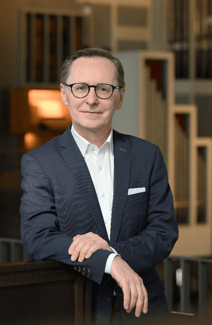 A man in a suit with glasses stands confidently, resting his hands on a surface, in an indoor setting.