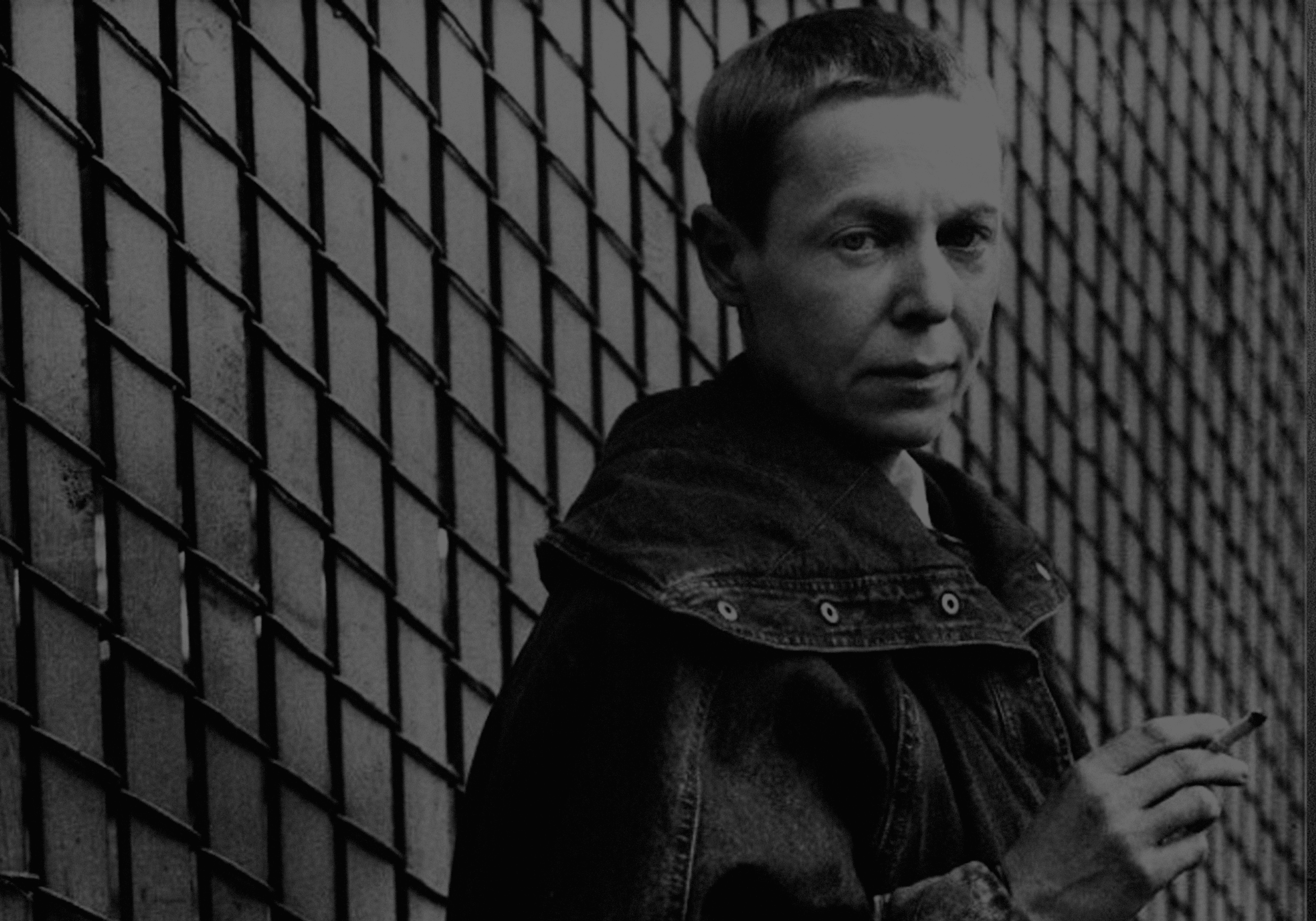 A person with short hair, wearing a jacket, holds a cigarette while standing next to a diamond-patterned wall.