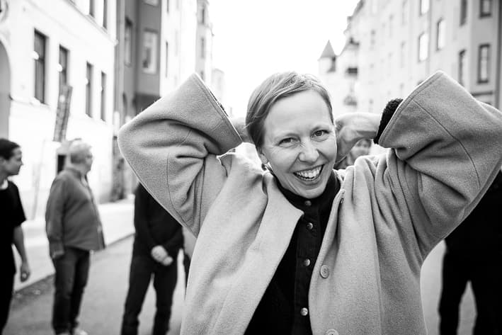 A person smiles while standing on a street with their hands raised behind their head; several other people are in the background.