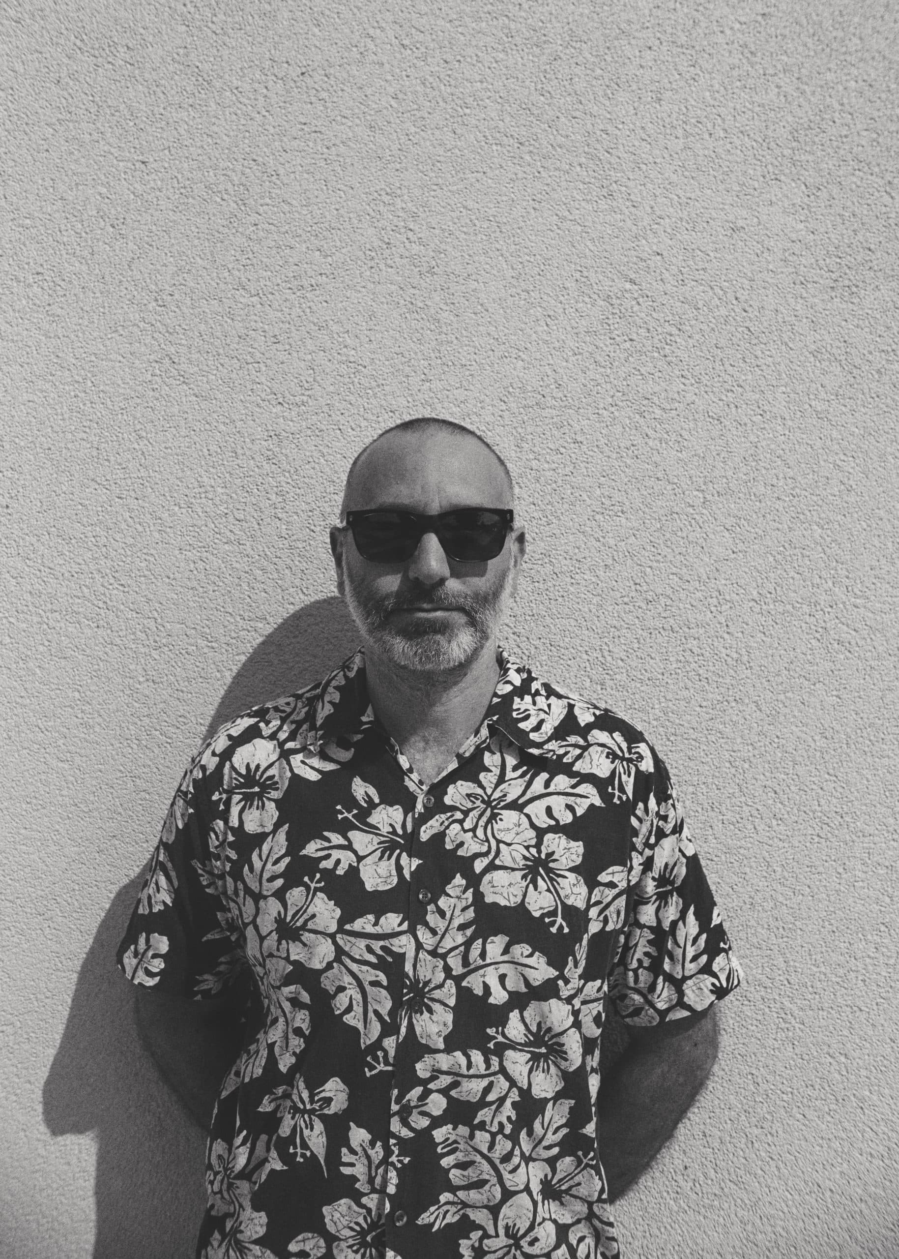 A man with a beard wearing sunglasses and a floral shirt stands against a textured wall.
