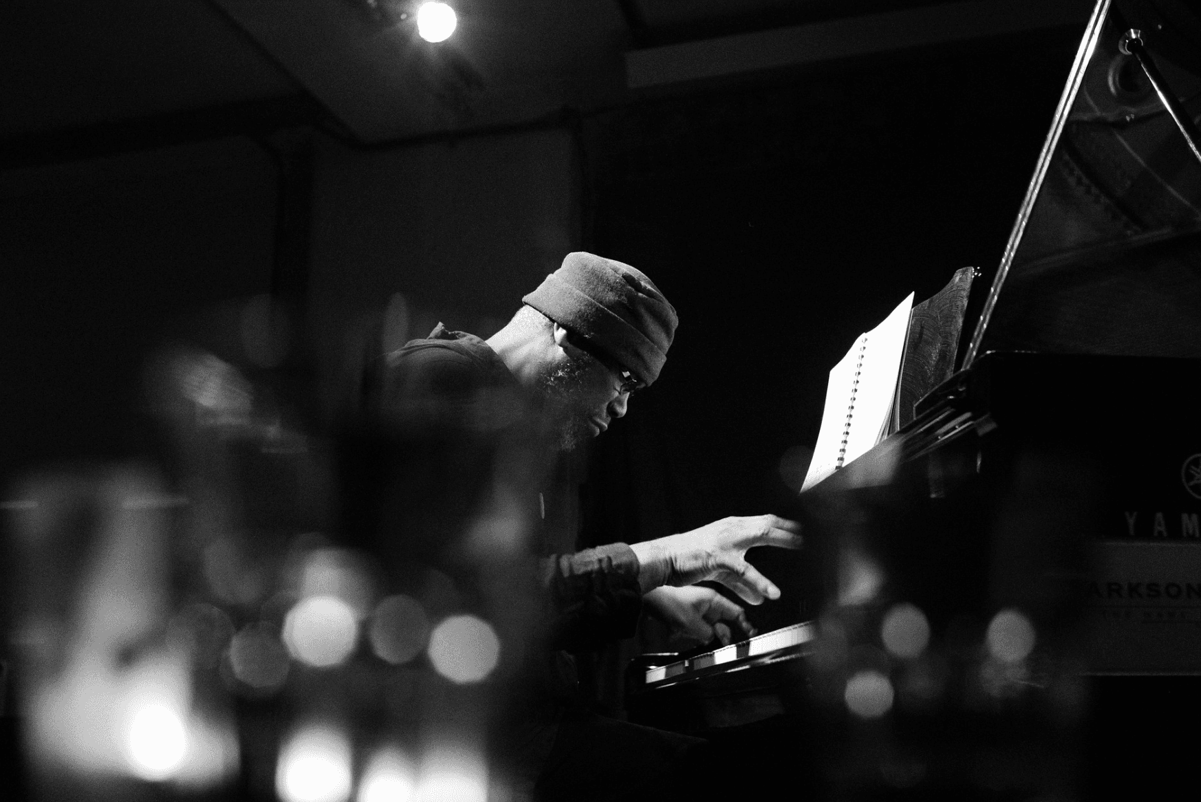 A person wearing a beanie is playing a piano with sheet music in a dimly lit room.