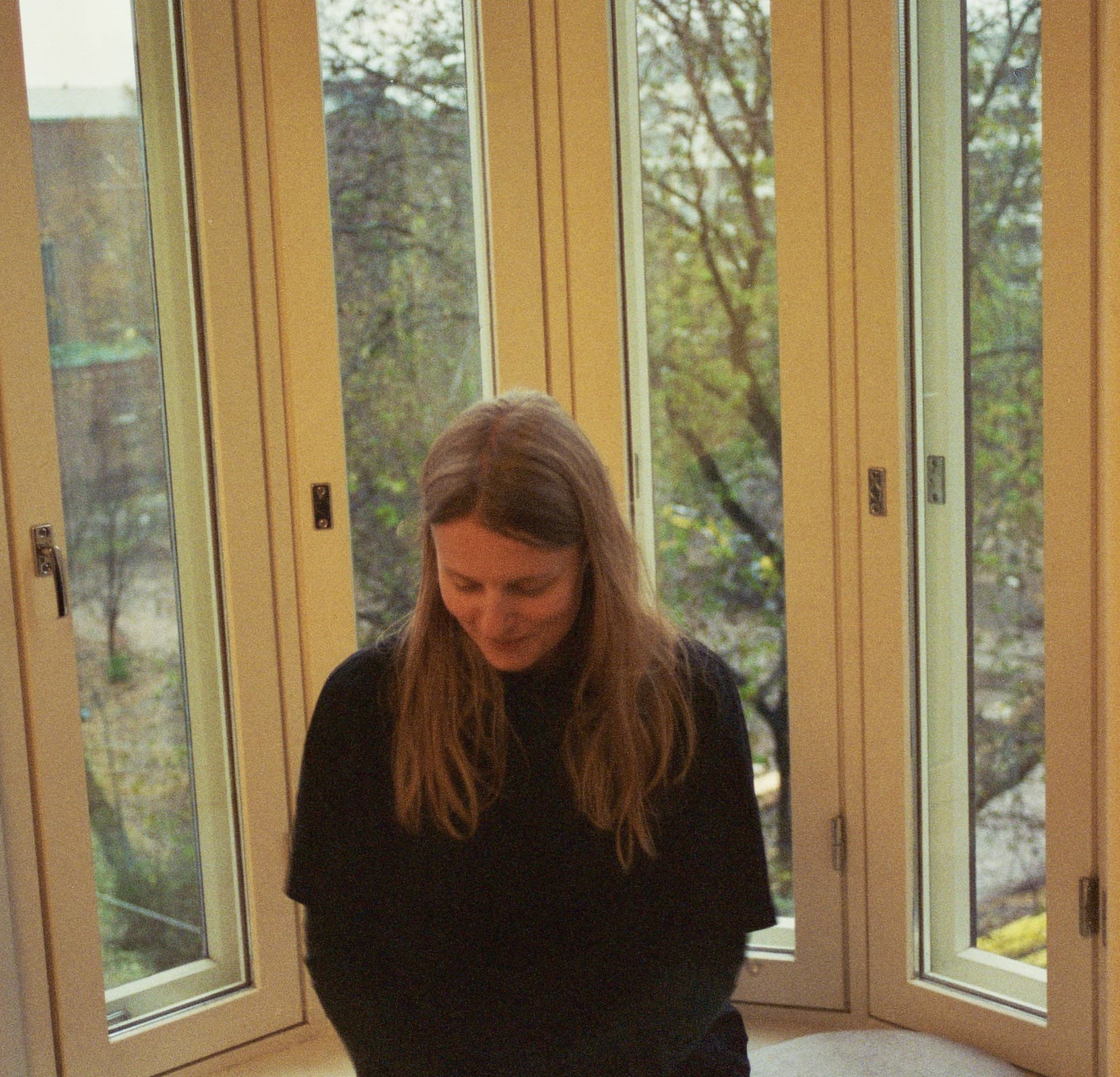 A person with long hair wearing a dark sweater is sitting by a large window, looking down.