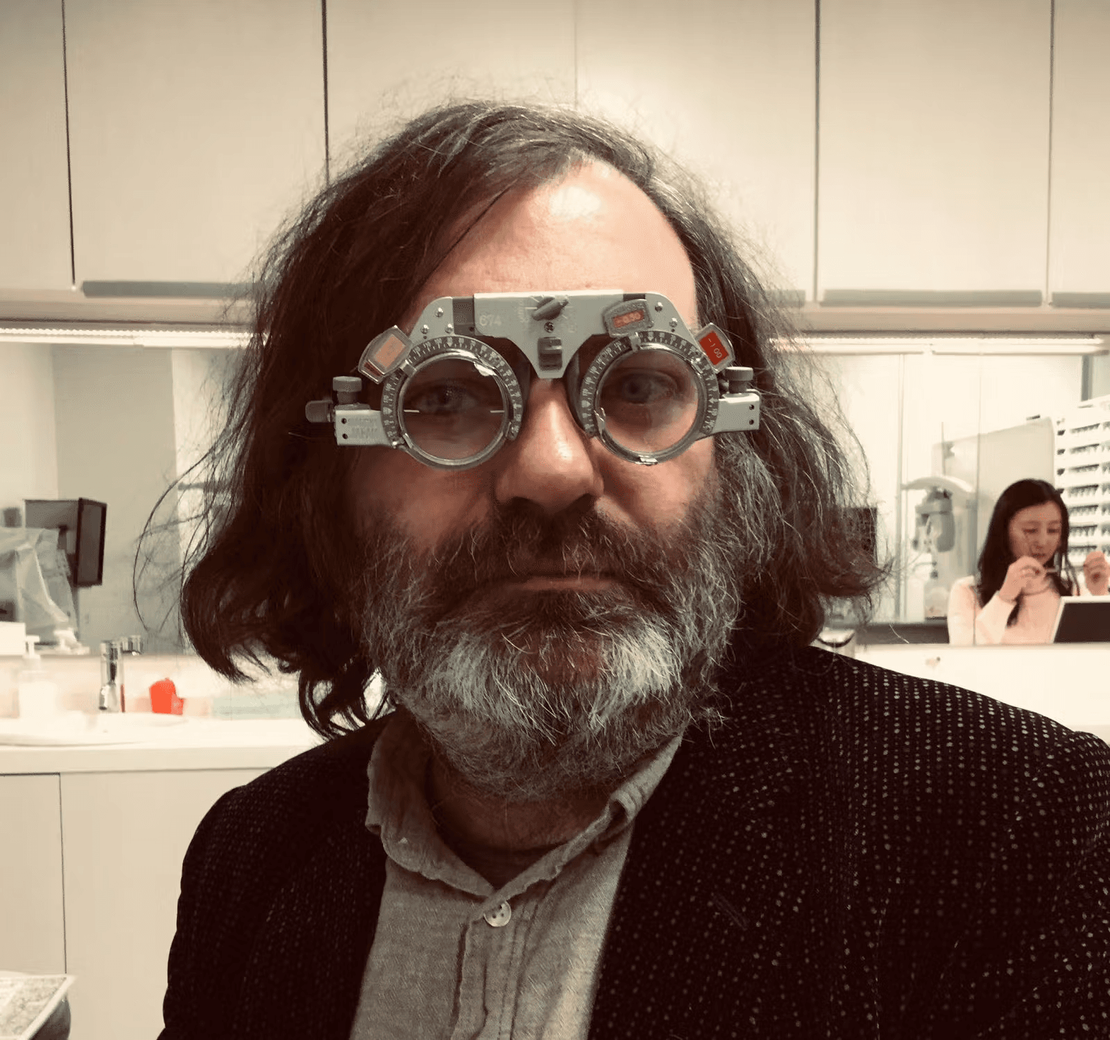 A man with long hair and a beard is wearing a trial lens frame in an optical examination room.