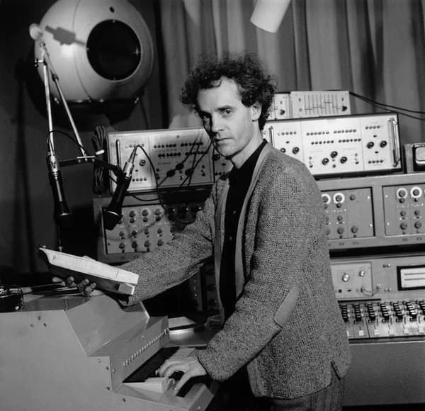 A man is standing at a synthesizer in a studio with various electronic equipment behind him.