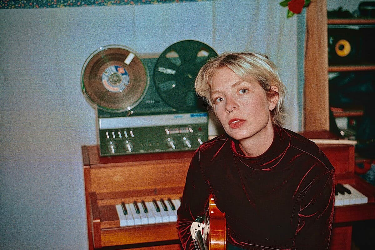 A person with short blonde hair holds a string instrument in front of a piano and a reel-to-reel tape recorder.