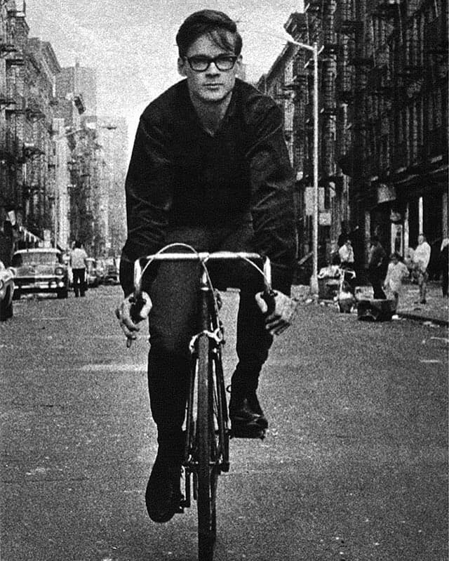 A person wearing glasses rides a bicycle down a city street, surrounded by buildings and pedestrians.