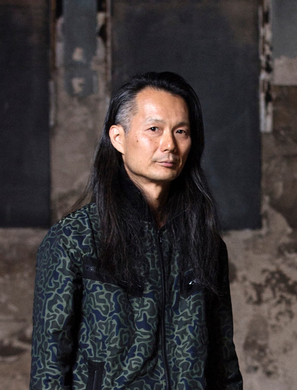 A person with long dark hair is wearing a patterned jacket, standing in front of a textured backdrop.