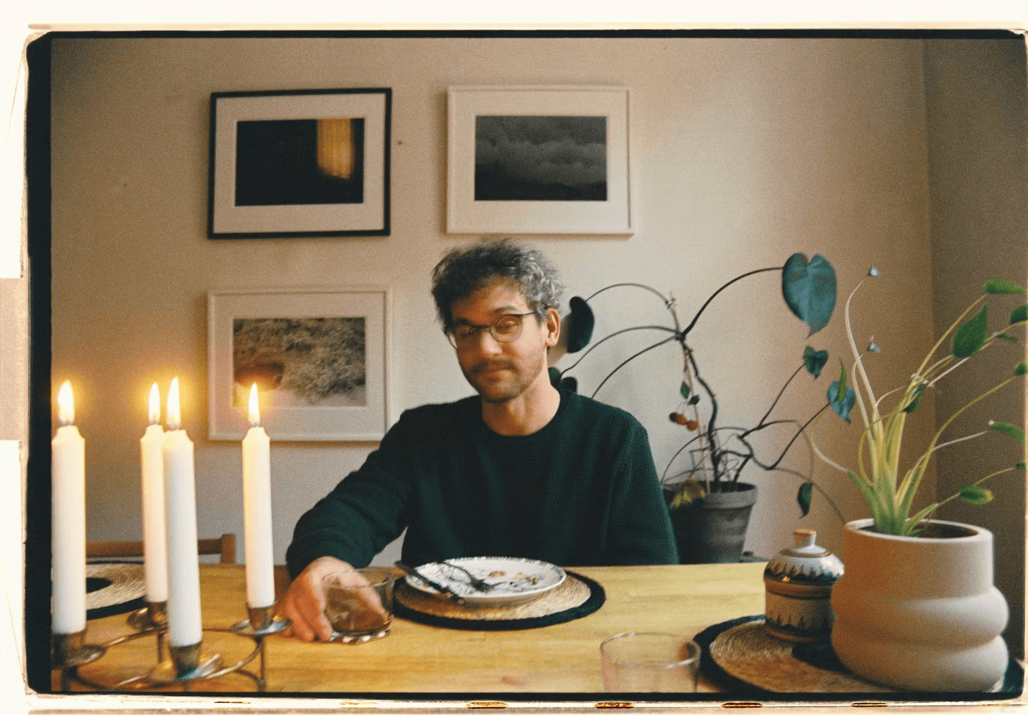 A person sits at a table with lit candles, plants, and framed pictures on the wall, holding a glass.