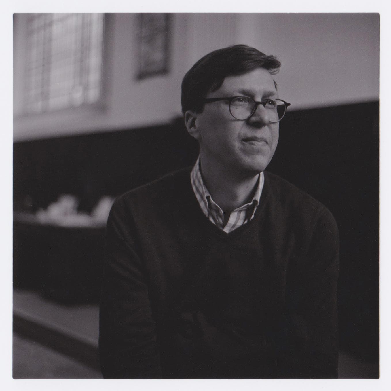 A man wearing glasses and a sweater sits indoors, looking to the side in a black and white photograph.