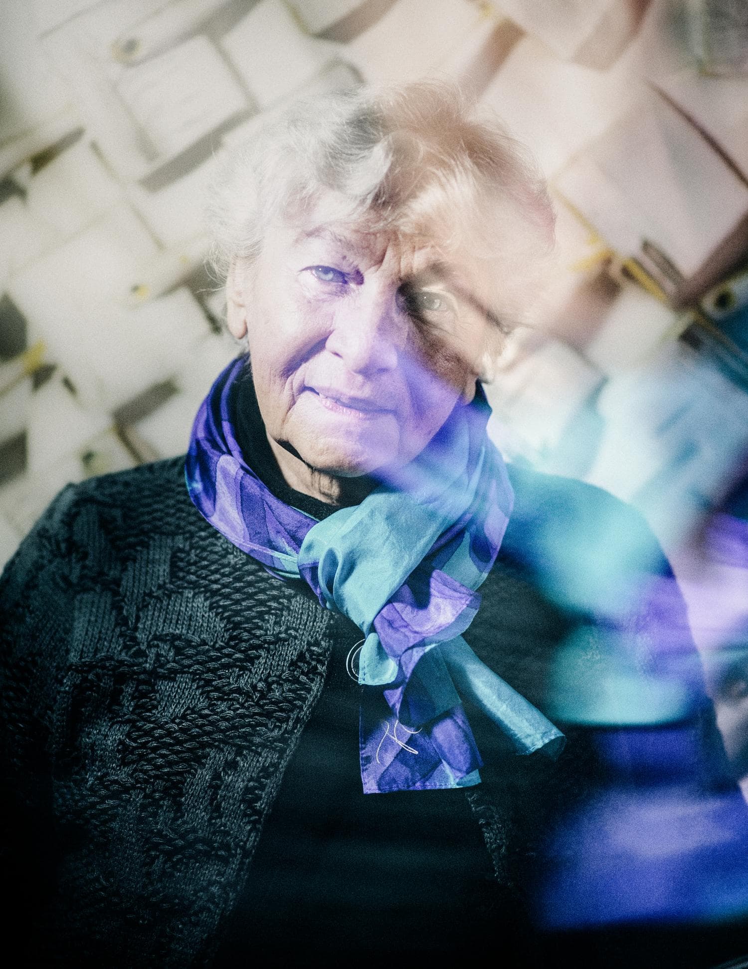 An elderly woman with gray hair and a colorful scarf stands against a backdrop of stacked paper, with a soft, dreamy overlay effect.