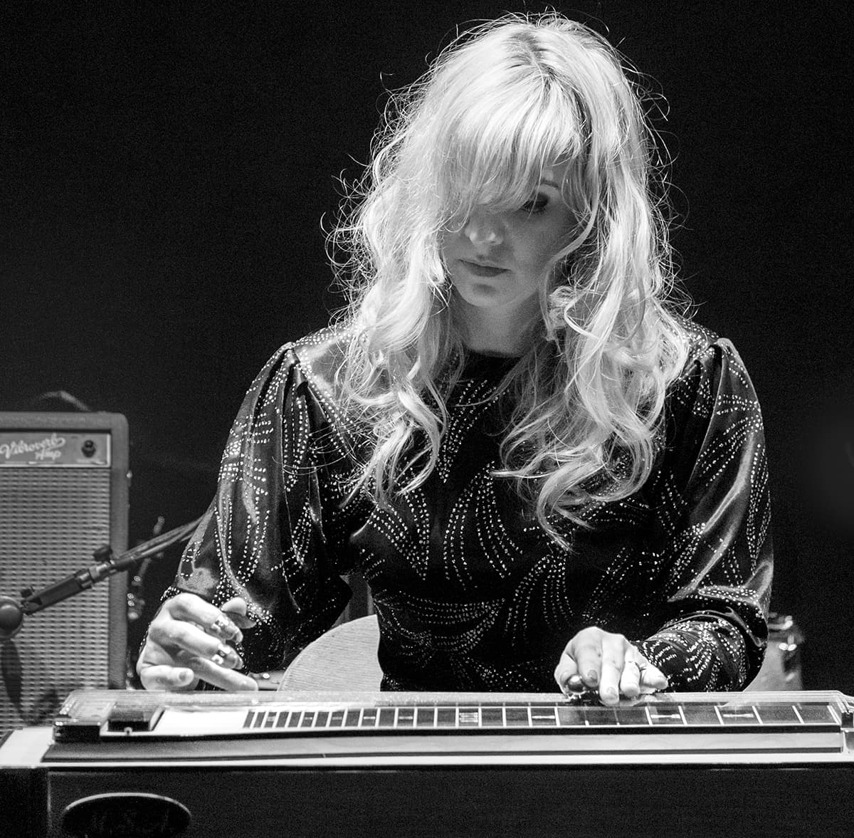 A person with long hair plays a pedal steel guitar on stage.