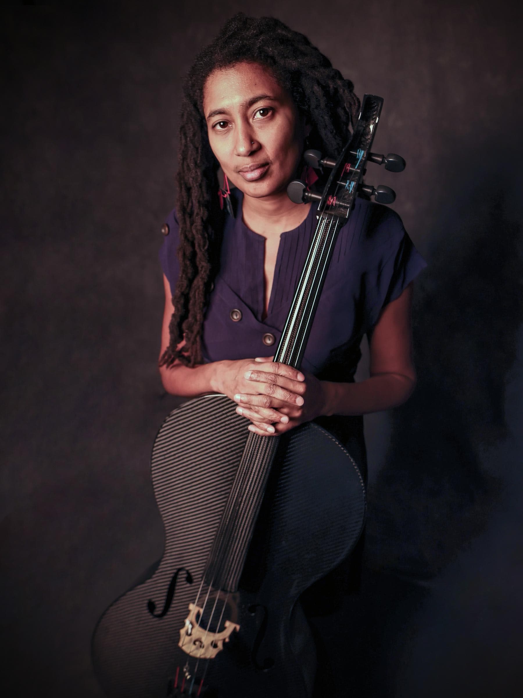 A person with long hair is holding a cello, wearing a dark dress, and standing against a plain background.