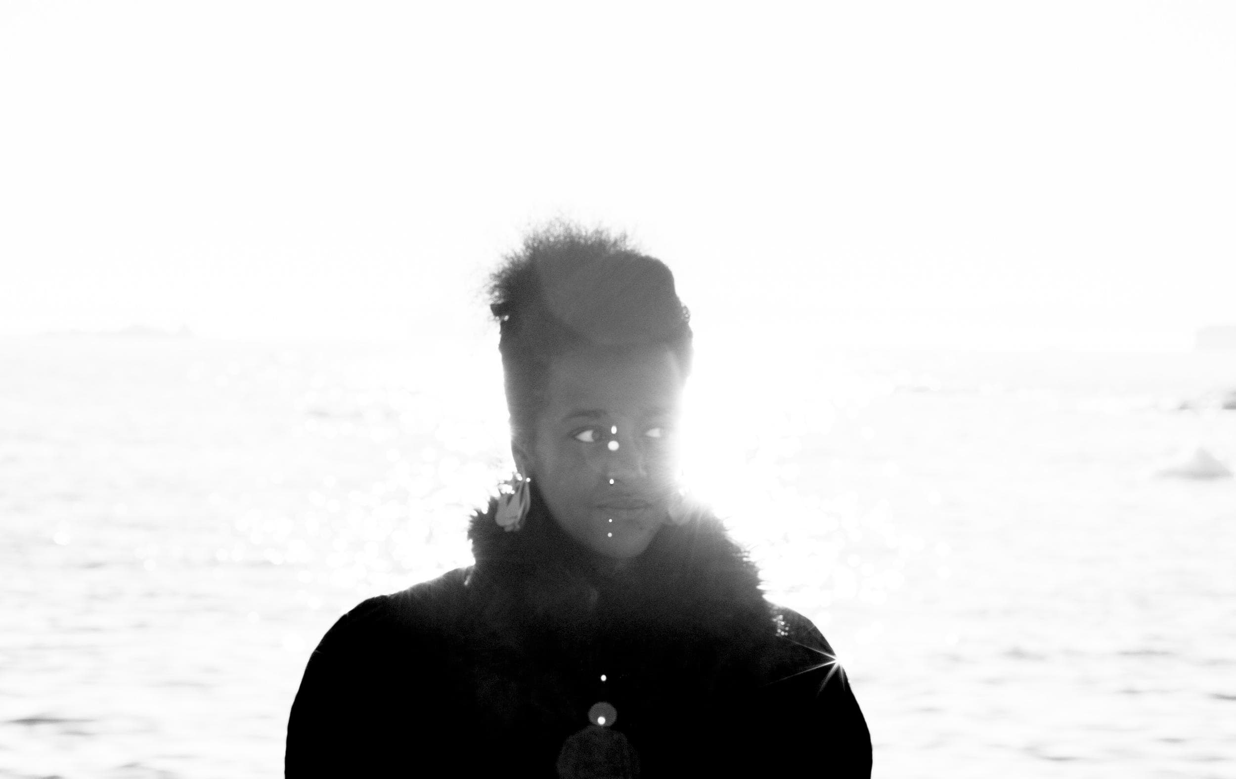 A person with a thoughtful expression stands near water, backlit by bright sunlight creating lens flare.