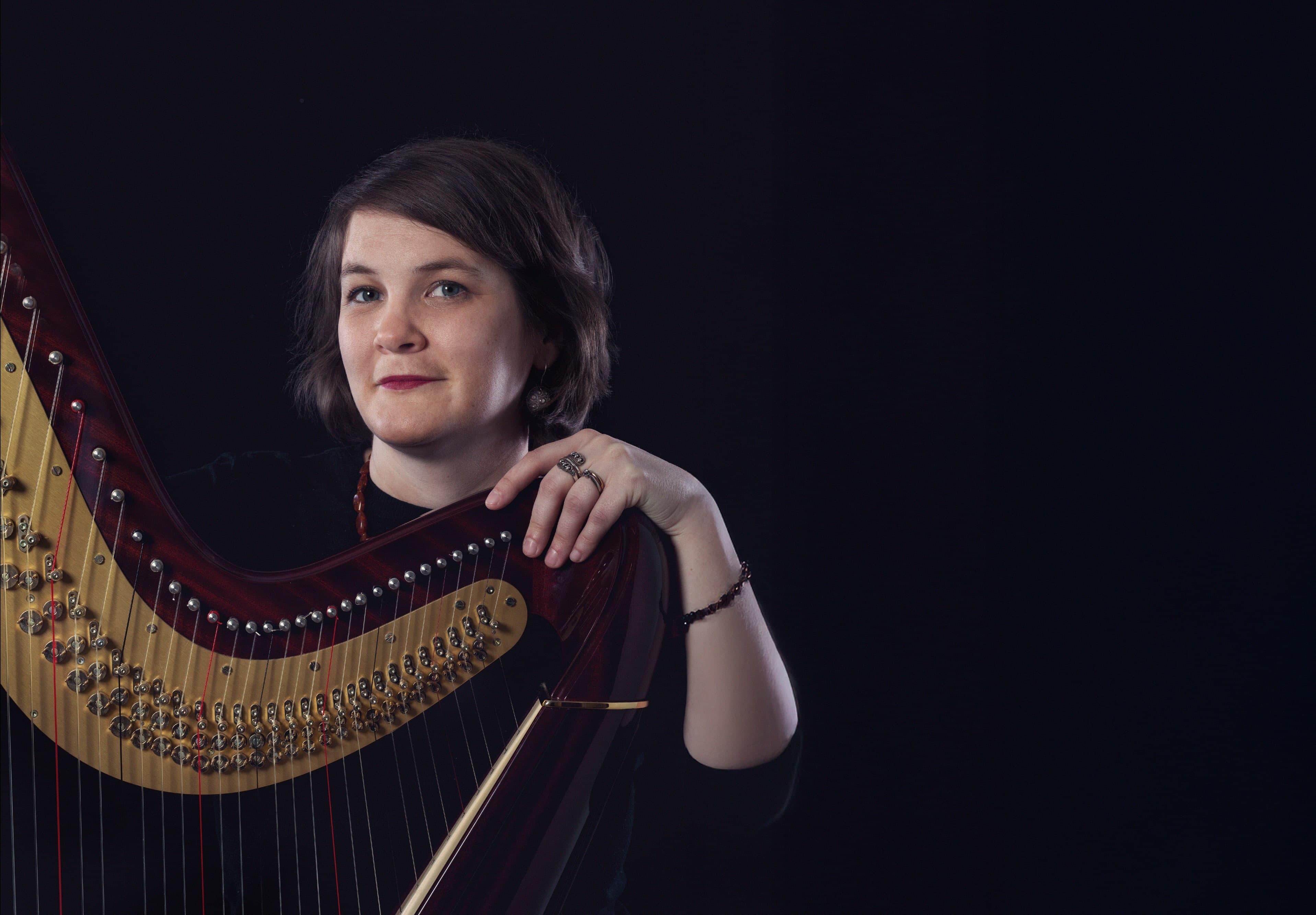 A person with short hair poses with a harp against a dark background.