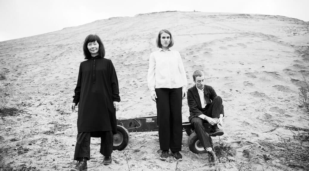 Three people are posing on a sandy landscape, with one sitting on a wagon while the other two stand beside it.