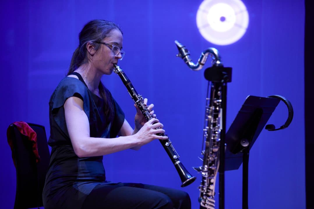 A woman is playing the clarinet on stage with a music stand in front of her, set against a blue-lit background.