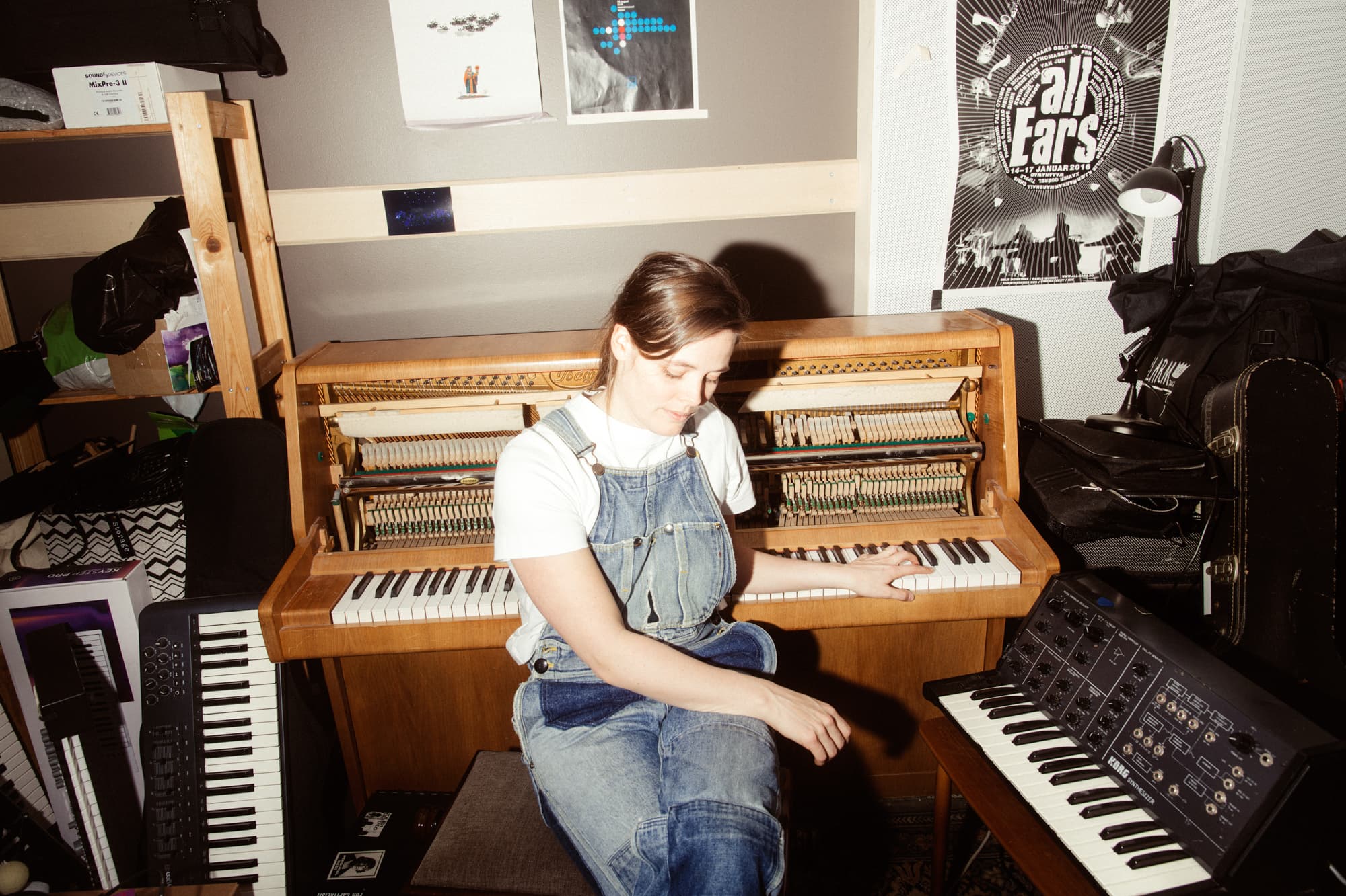 A person in overalls sits at a wooden piano in a cluttered room with keyboards and posters on the walls.