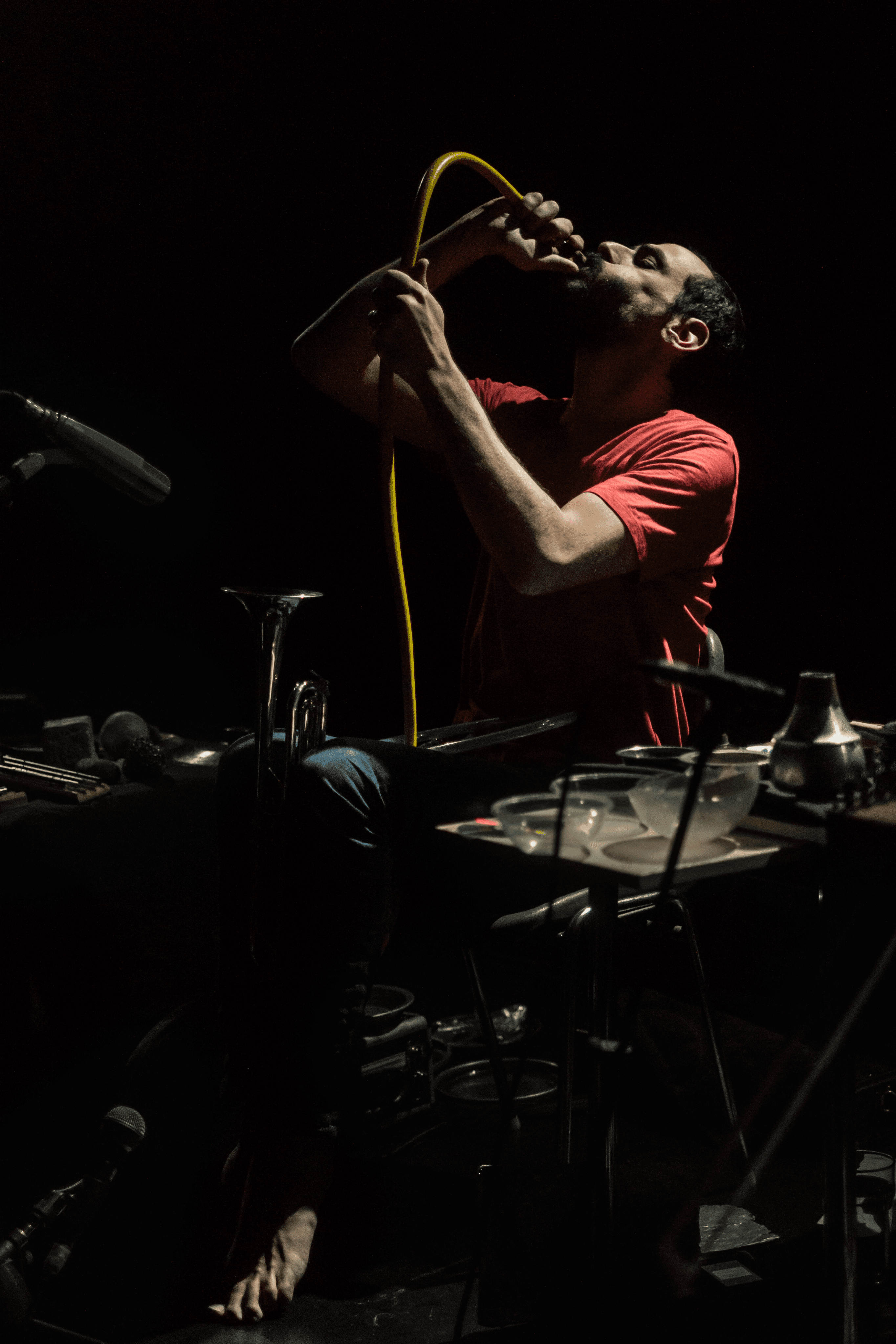 A person in a red shirt is performing with a yellow tube in their mouth, surrounded by musical instruments and equipment.
