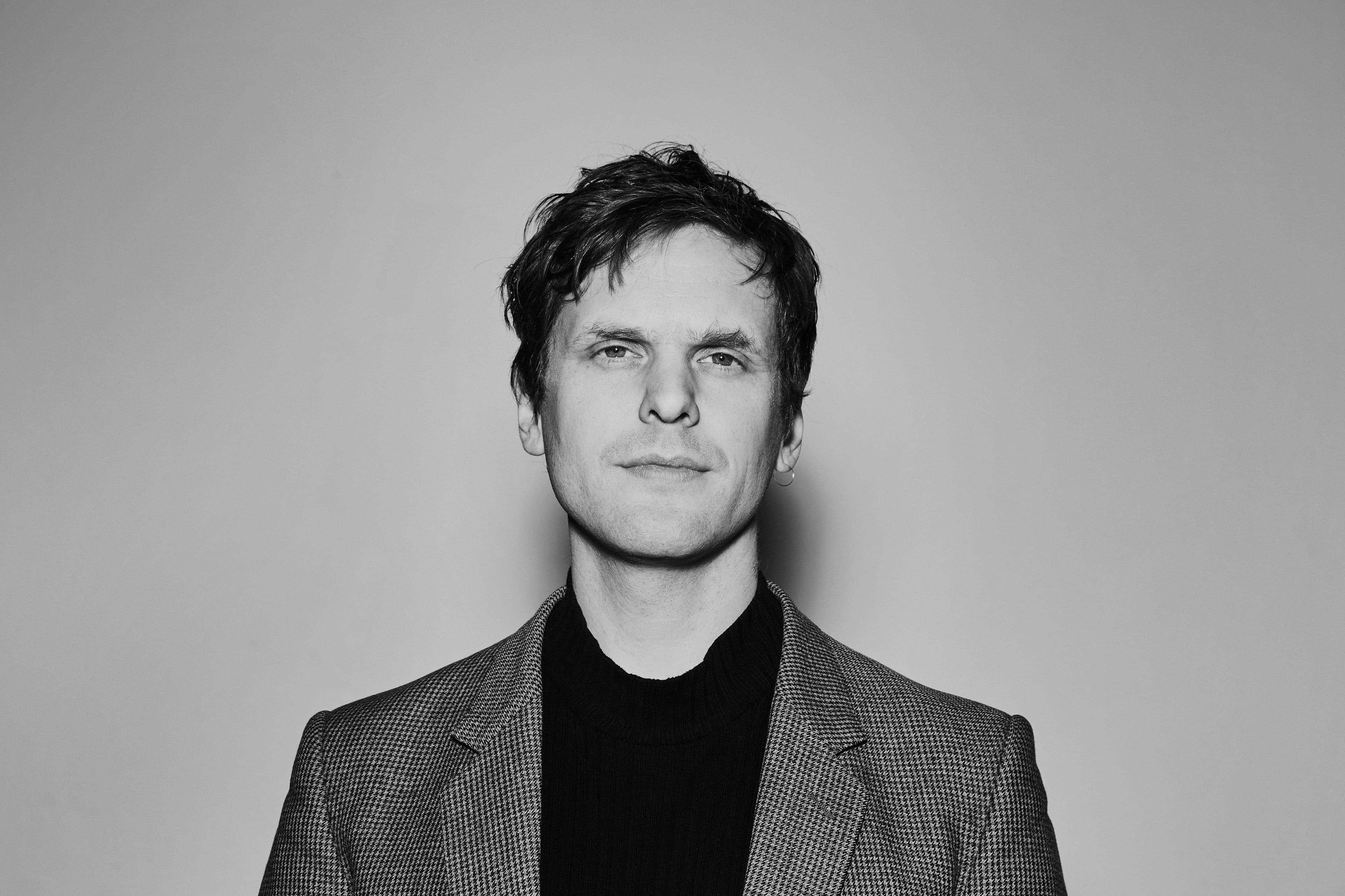 A man with short hair wearing a checkered blazer and a dark shirt, photographed in black and white against a plain background.