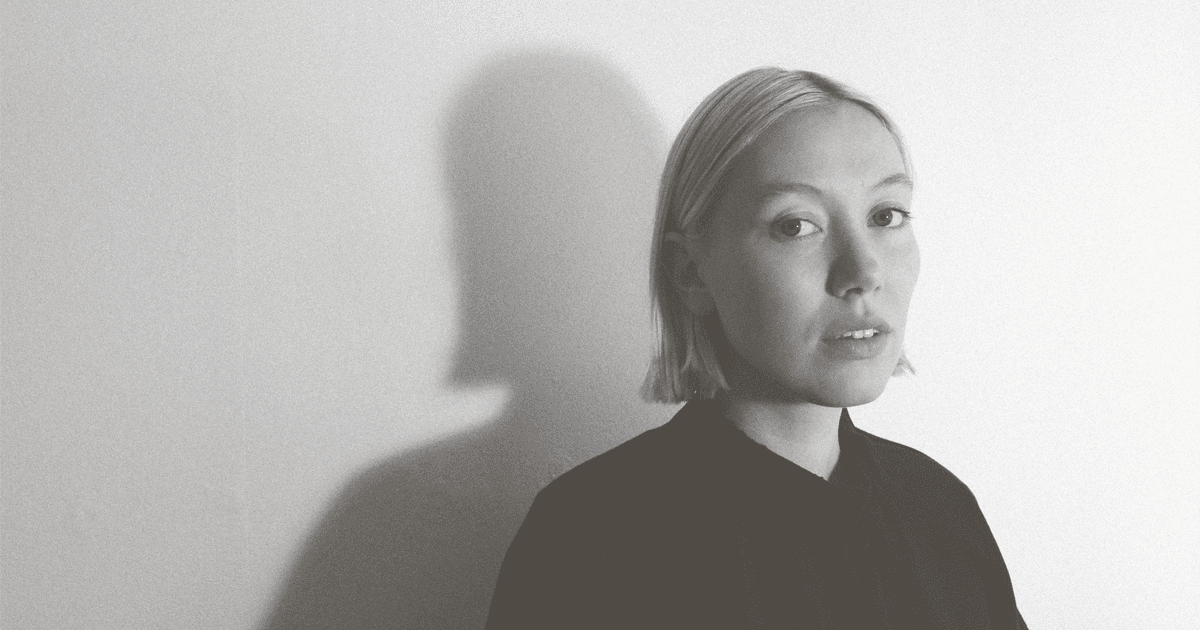 A woman with short hair is looking at the camera in a black and white setting, casting a shadow on a plain wall.