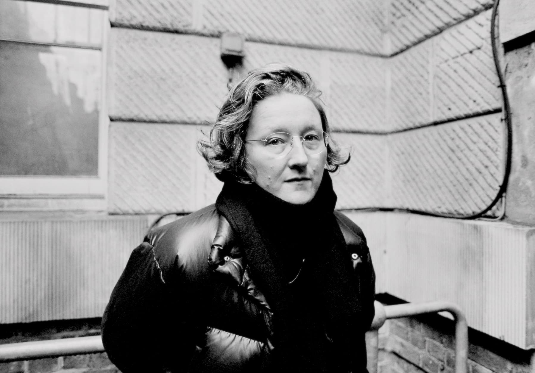 A person wearing glasses and a puffy jacket stands against an outdoor wall.