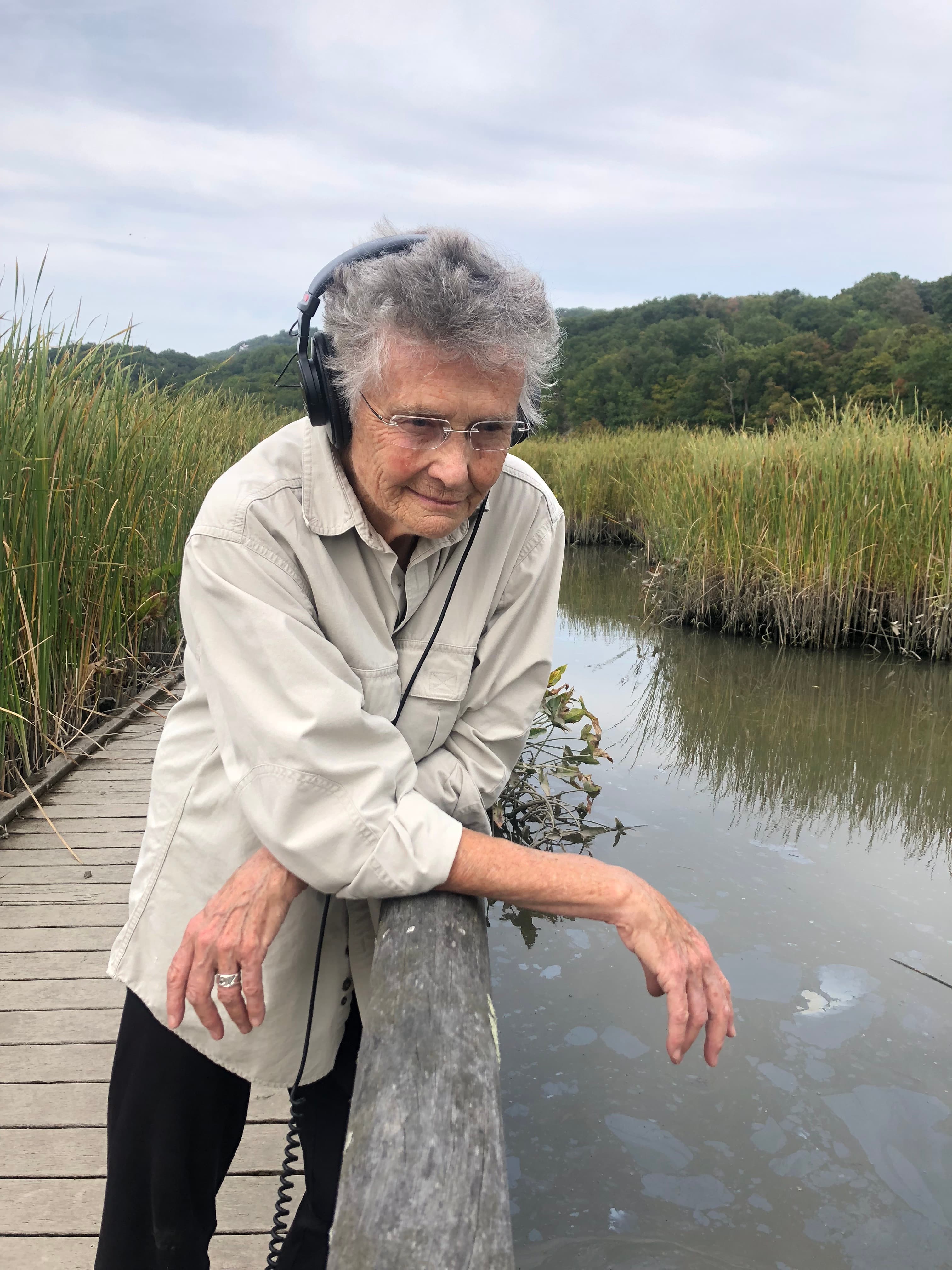 An elderly person wearing headphones leans on a wooden railing by a marshy area with tall grasses and a calm waterway.
