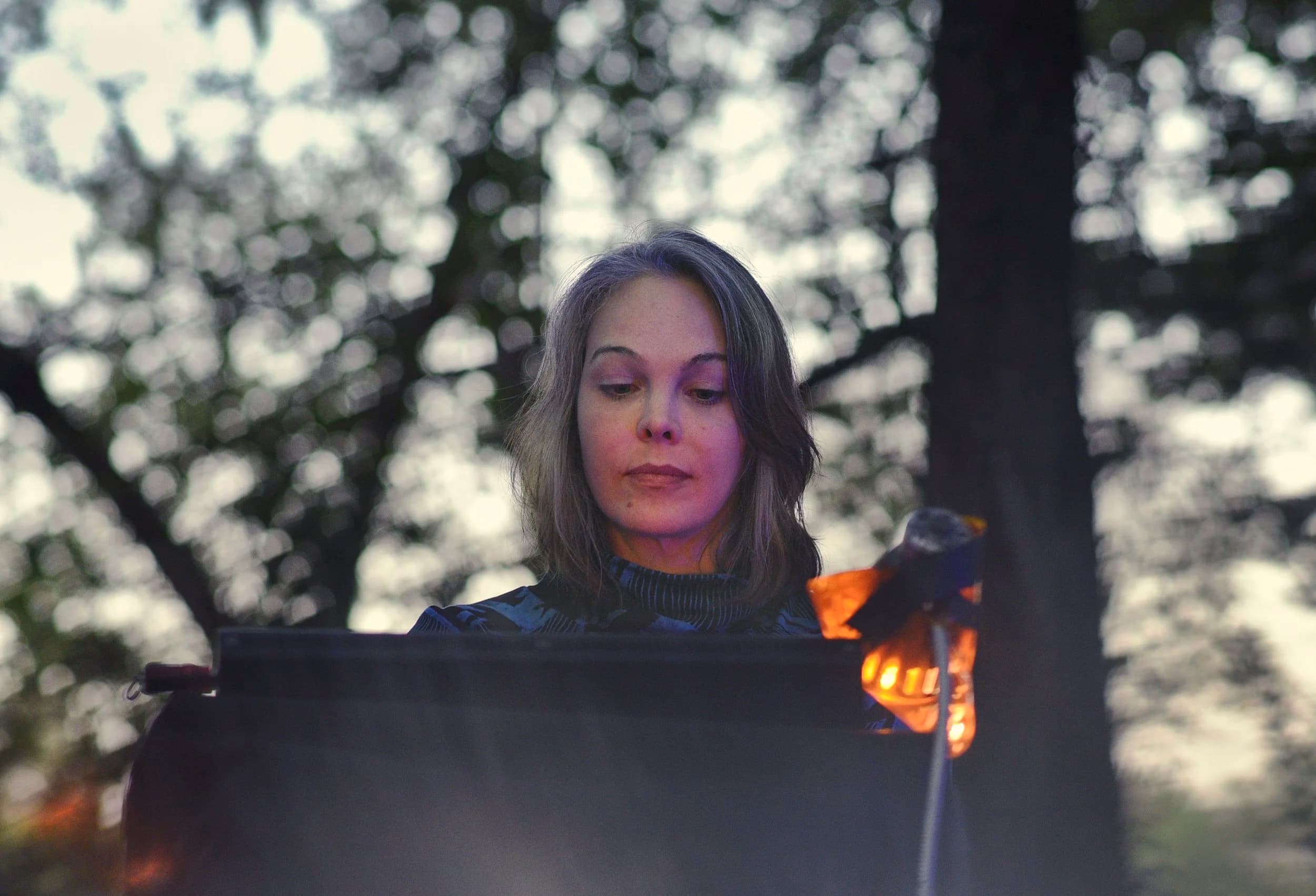 A woman stands outdoors in front of a music stand, with trees blurred in the background.
