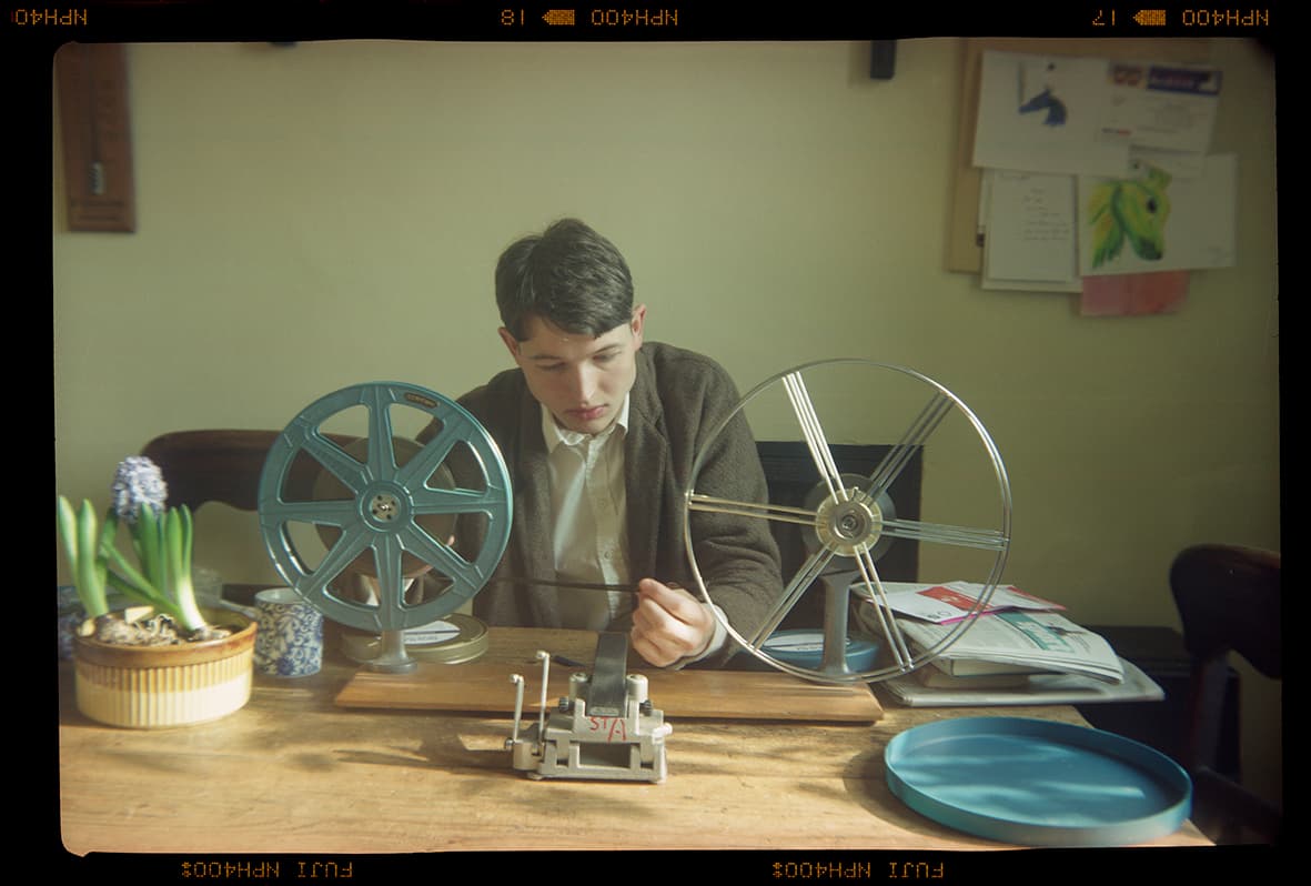 A person sits at a table examining film reels, with a flower pot and some books nearby.