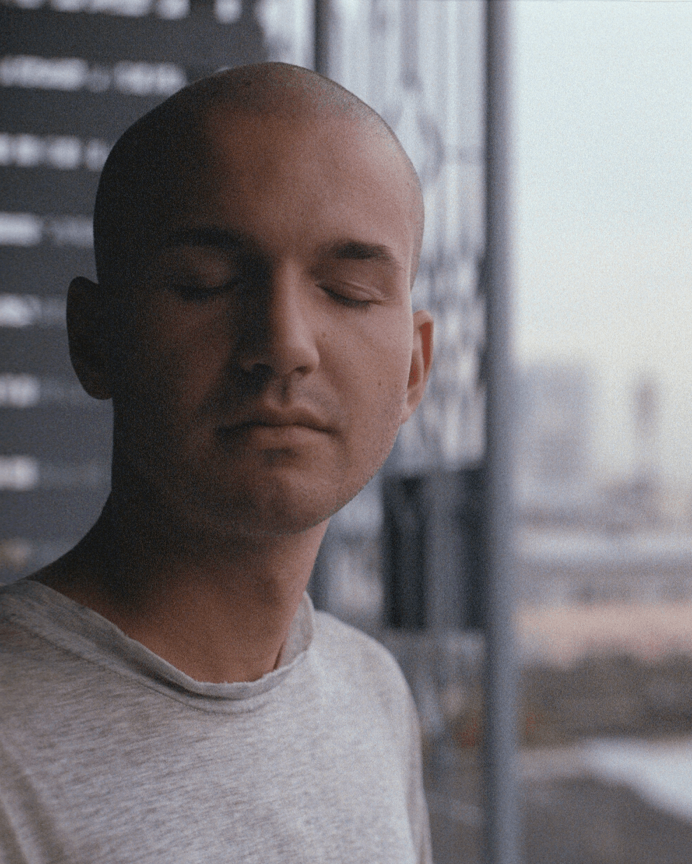 A person with closed eyes is standing outdoors, wearing a light gray shirt, with a blurred background.