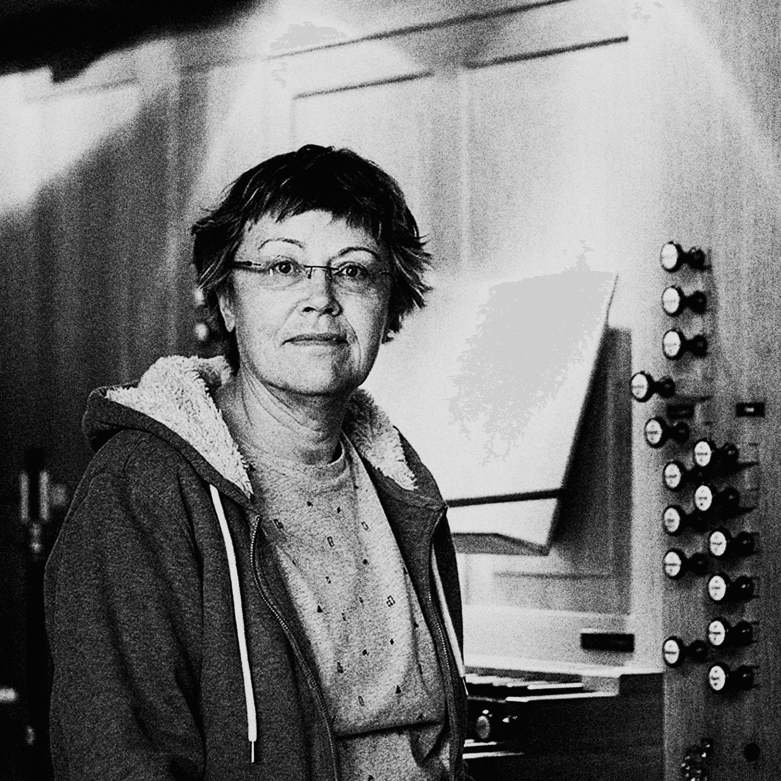 A person wearing glasses and a hoodie is standing in a room with paneling and a control panel in the background.