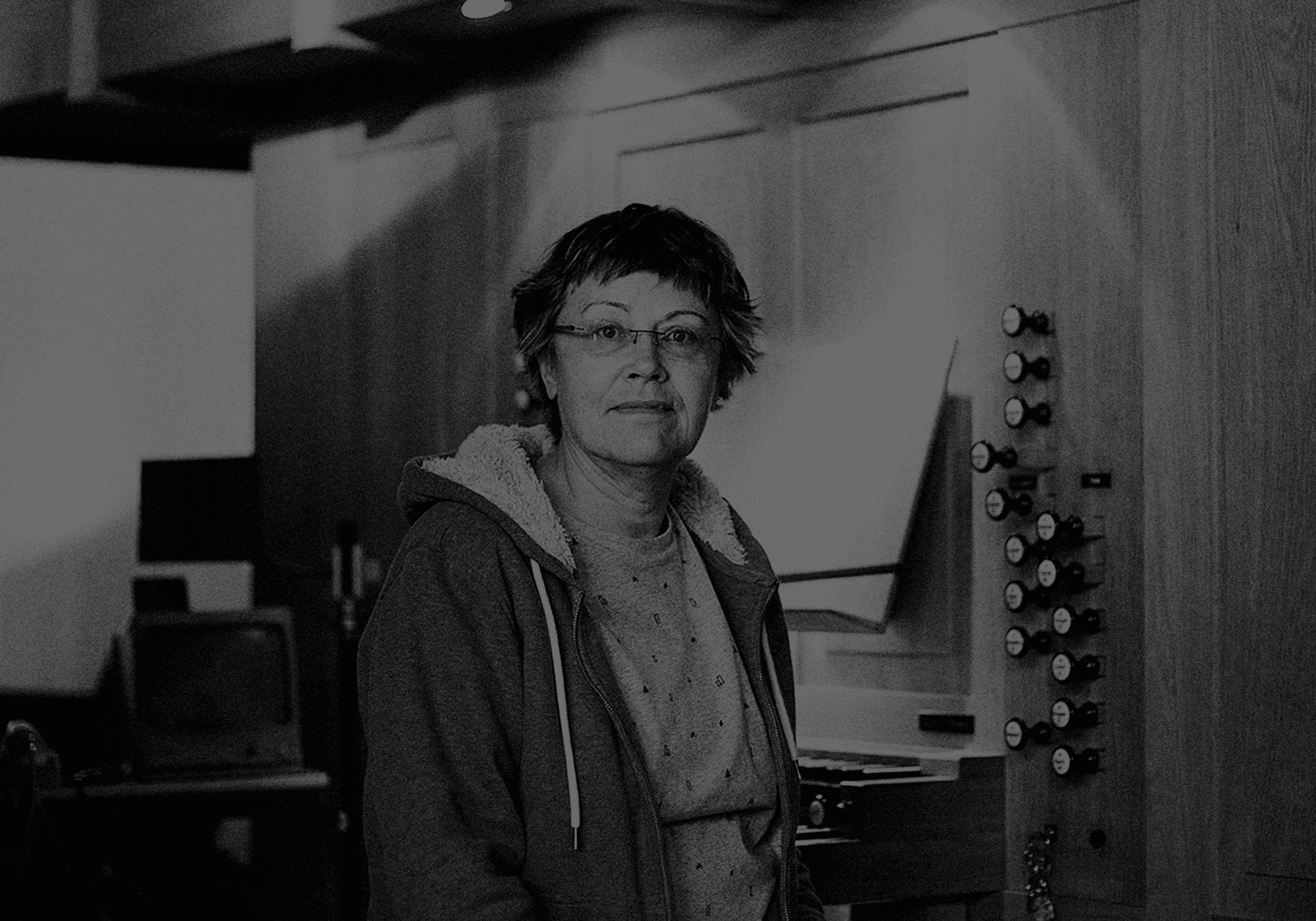 A person wearing glasses and a hoodie stands in a room with wood paneling and various buttons or knobs on the wall.