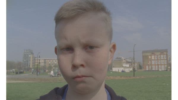 A young boy with a concerned expression stands in a park with buildings in the background.