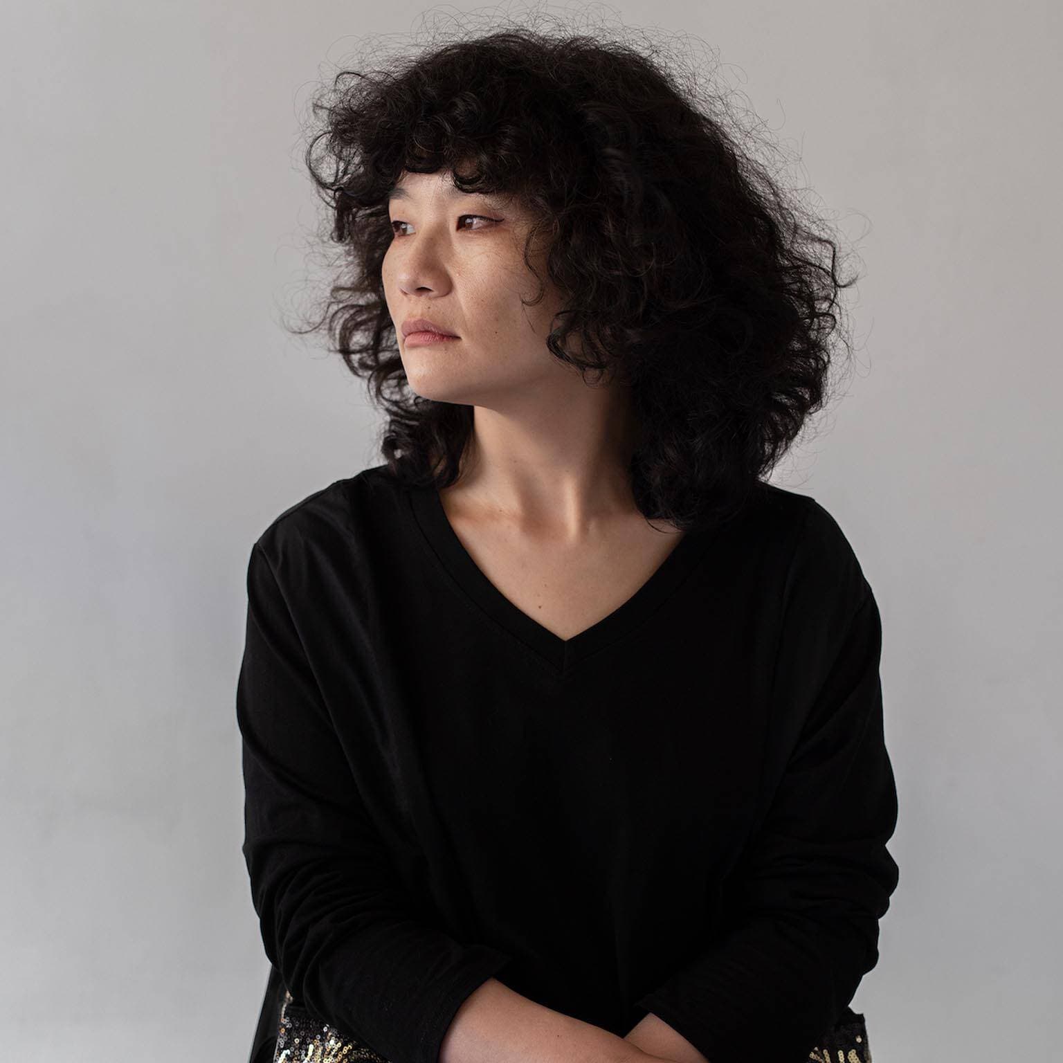 A person with curly hair in a black shirt looks to the side against a plain backdrop.